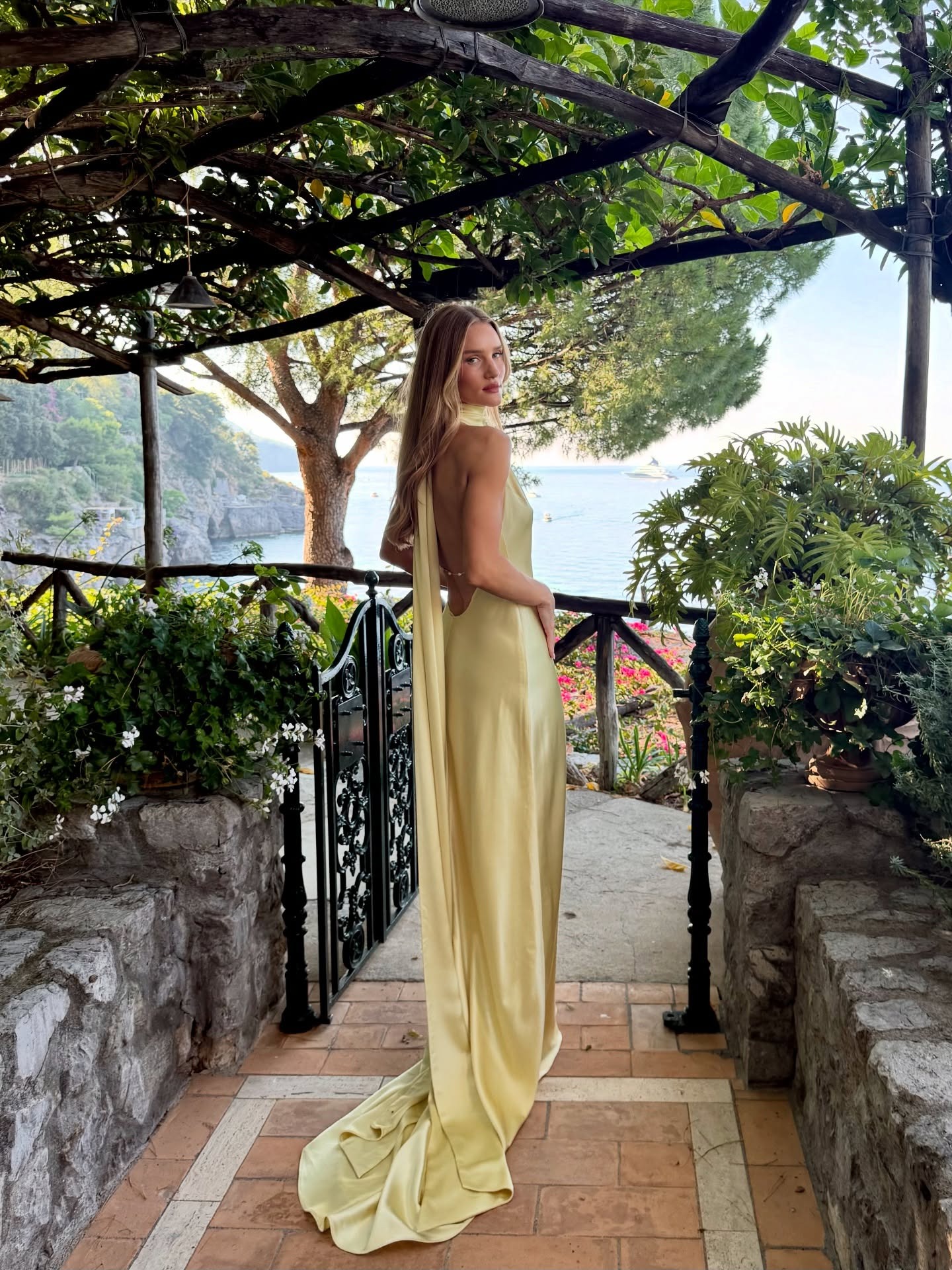 Model @rosiehw stands outside on a balcony in Italy wearing a satin maxi dress in butter yellow.