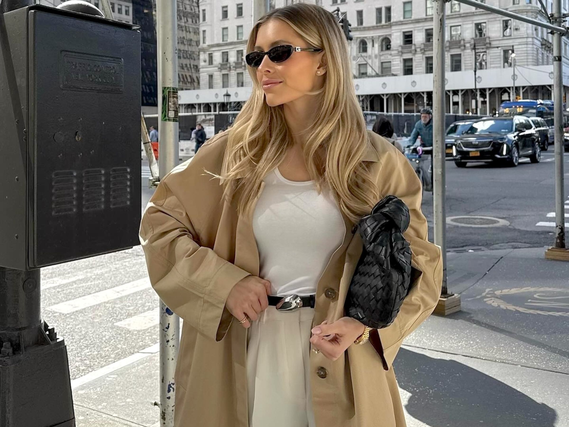 influencer Fredrika Ekerot poses on a NYC sidewalk wearing black oval sunglasses, a white tank top, oversized trench coat, black belt with a silver buckle, a black woven bag, and tan pants