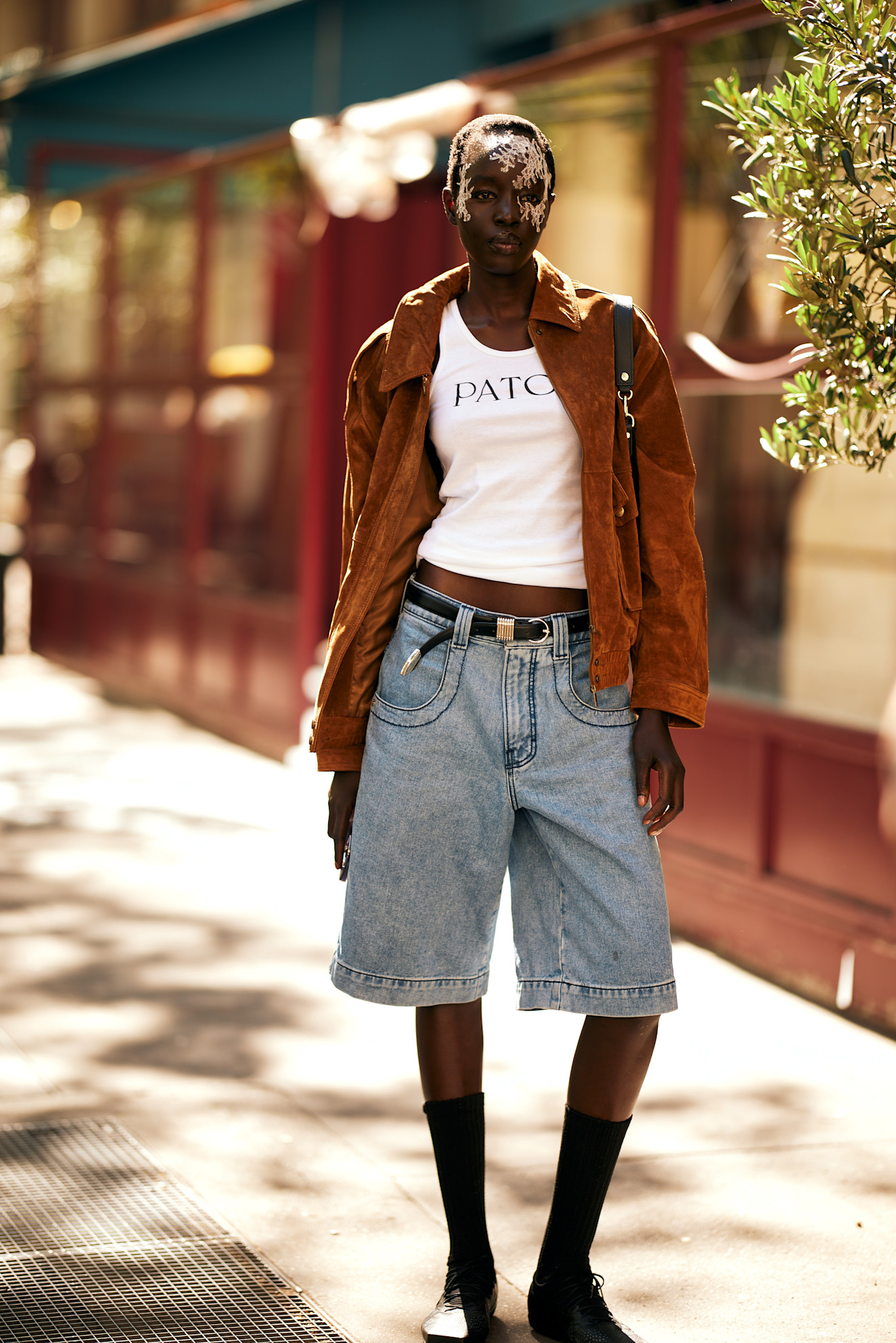 A model outside of a show in Paris wearing a suede jacket, white Patou tank top, and Bermuda jean shorts.