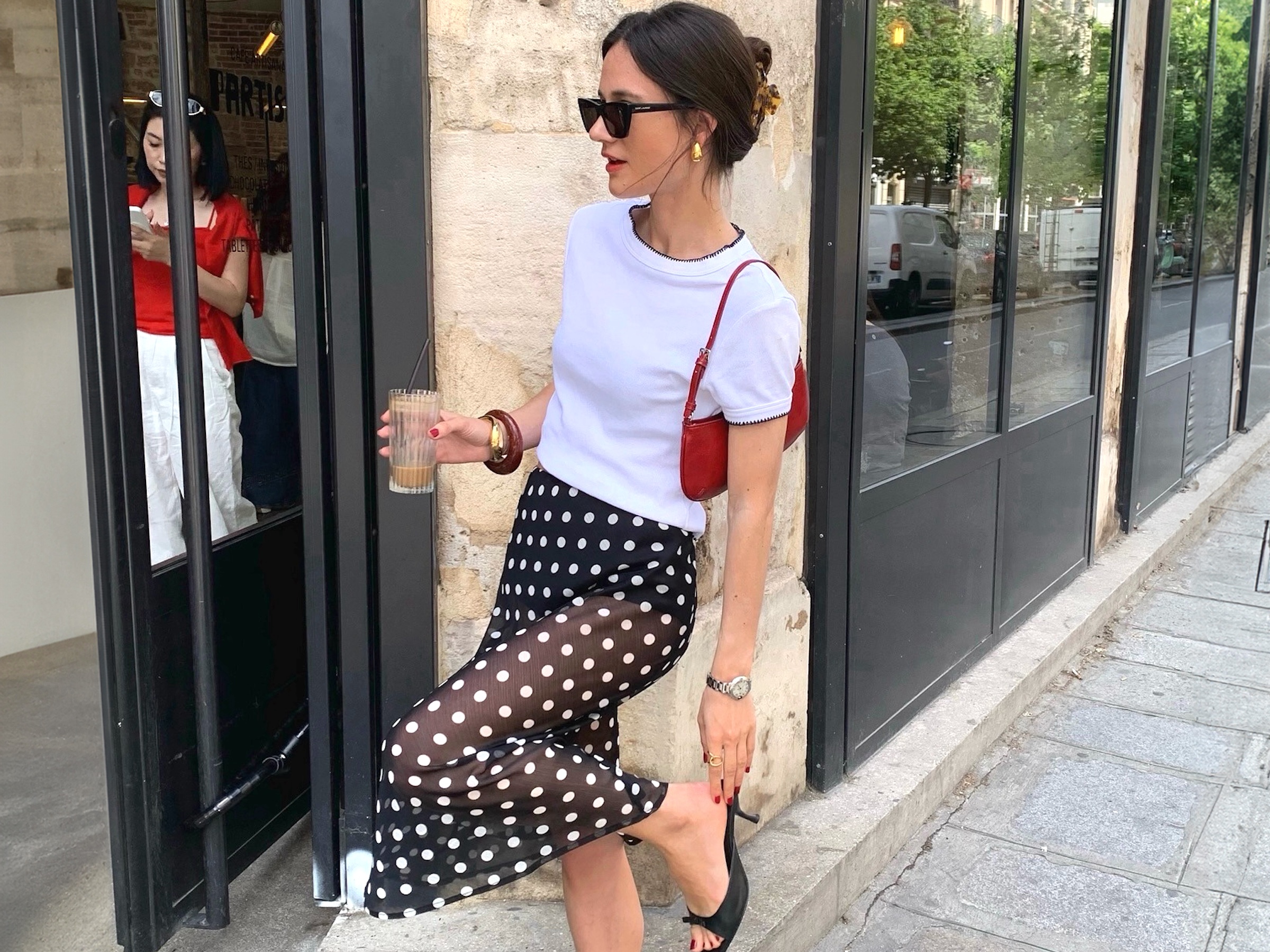 French style influencer Leasy In Paris poses outside a cafe in Paris holding a ribbed glass while wearing a claw hair clip, black sunglasses, yellow-gold chunky earrings, a white contrast hem T-shirt, mini red shoulder bag, bangles, a polka-dot skirt, simple silver watch, and black kitten-heel mule sandals
