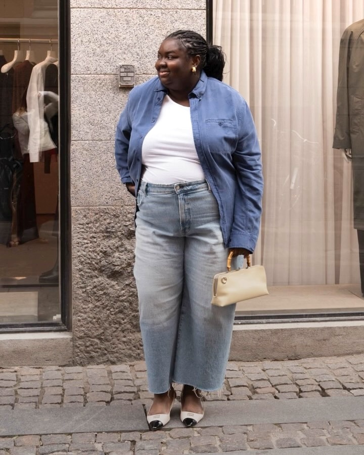 British plus size style influencer Abisola Omole poses on a sidewalk during Copenhagen Fashion Week wearing a blue button-down shirt, white basic top, faded Madewell straight-leg jeans with raw hems, and pointed cap-toe Miu Miu slingback kitten heels, and a bamboo top-handle mini bag