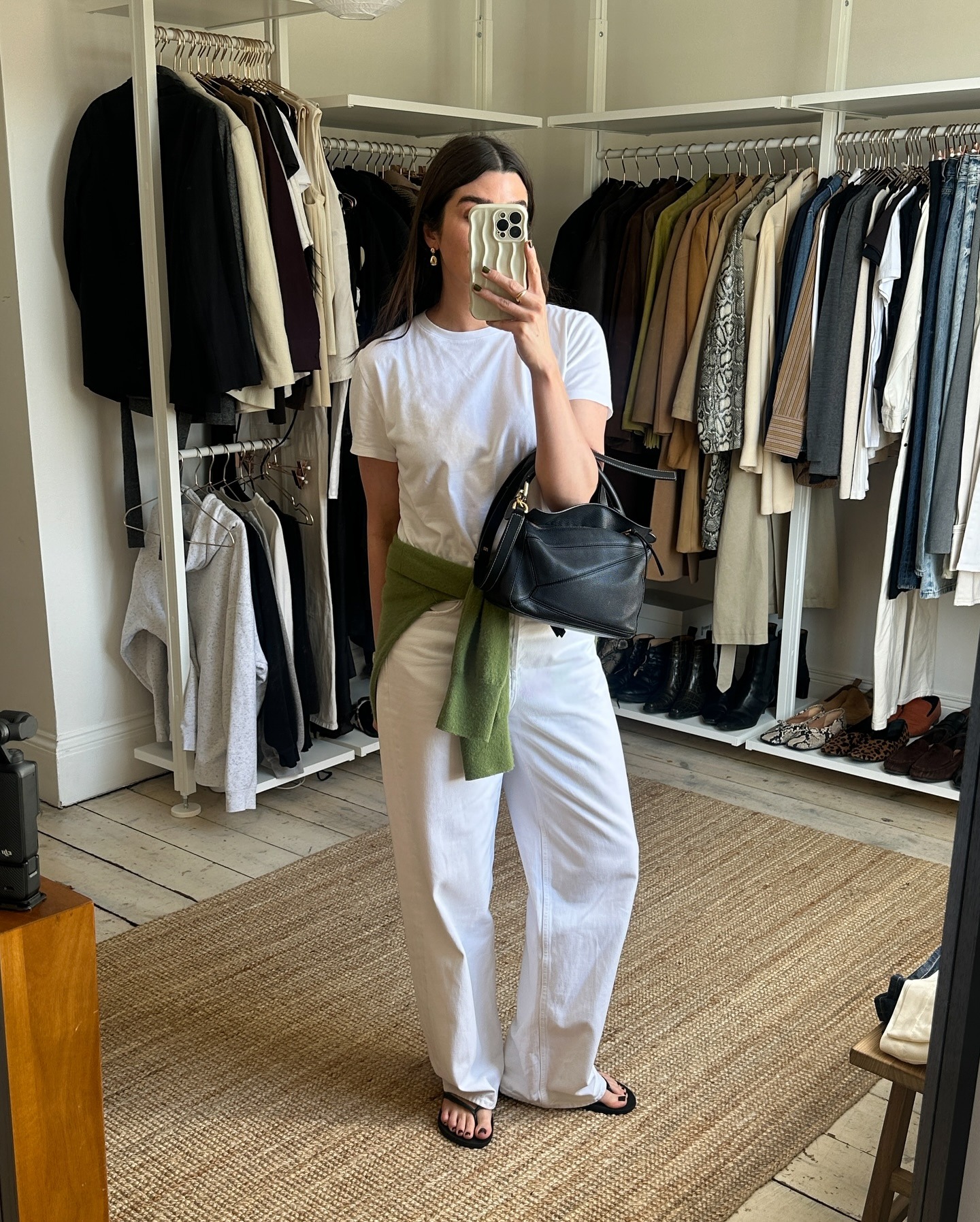British fashion influencer Anna Newton poses for a mirror selfie in her cool London closet wearing a white T-shirt, green sweater tied around her waist, a black Loewe puzzle bag, relaxed white jeans, and black flat flip-flop sandals