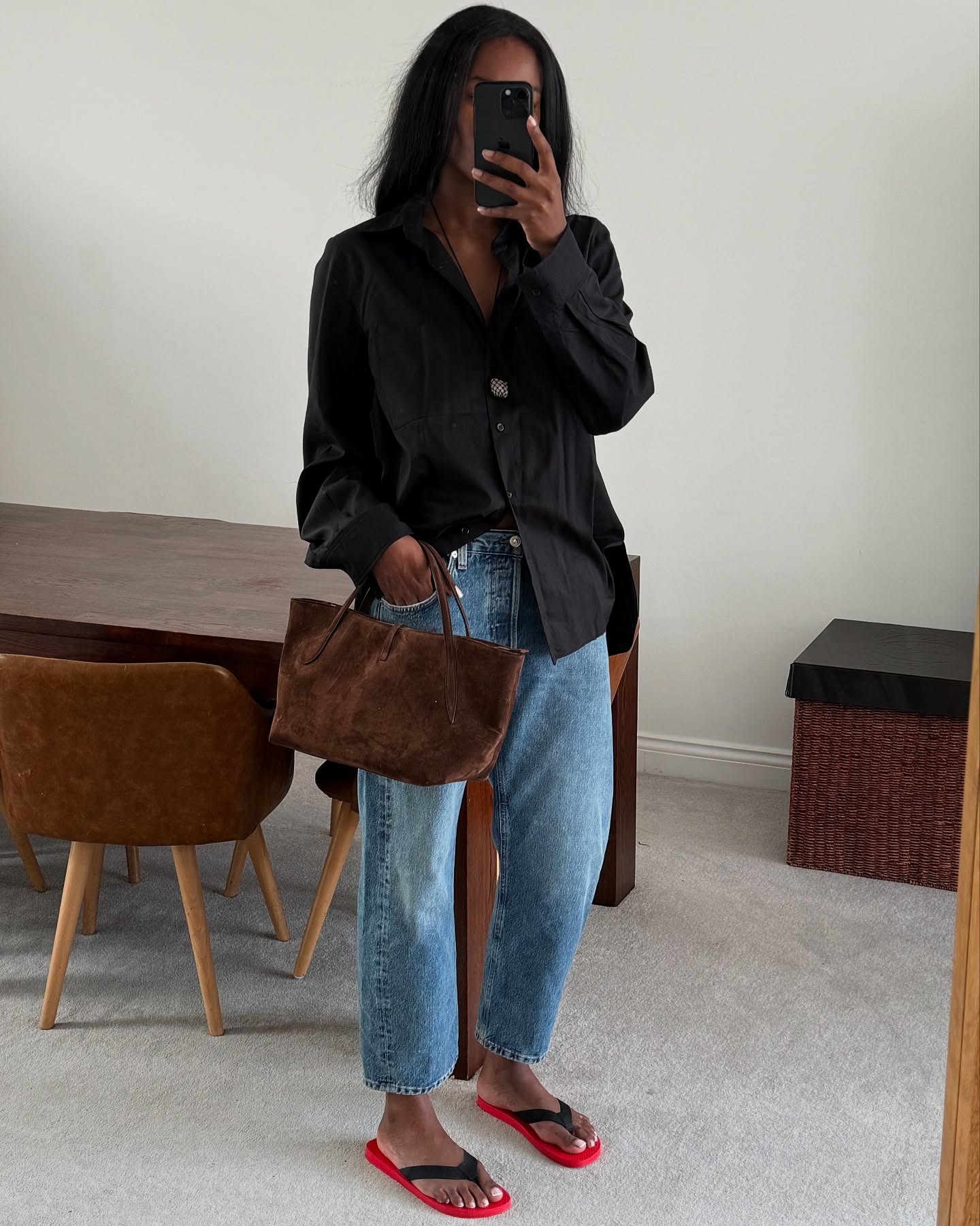 British style influencer Marilyn Nwawulor-Kazemaks poses for a mirror selfie in her London living room wearing a black linen button-down shirt, shell pendant necklace, cropped ankle-length straight-leg jeans, a small brown suede tote bag, and The Row black and red flip-flop sandals