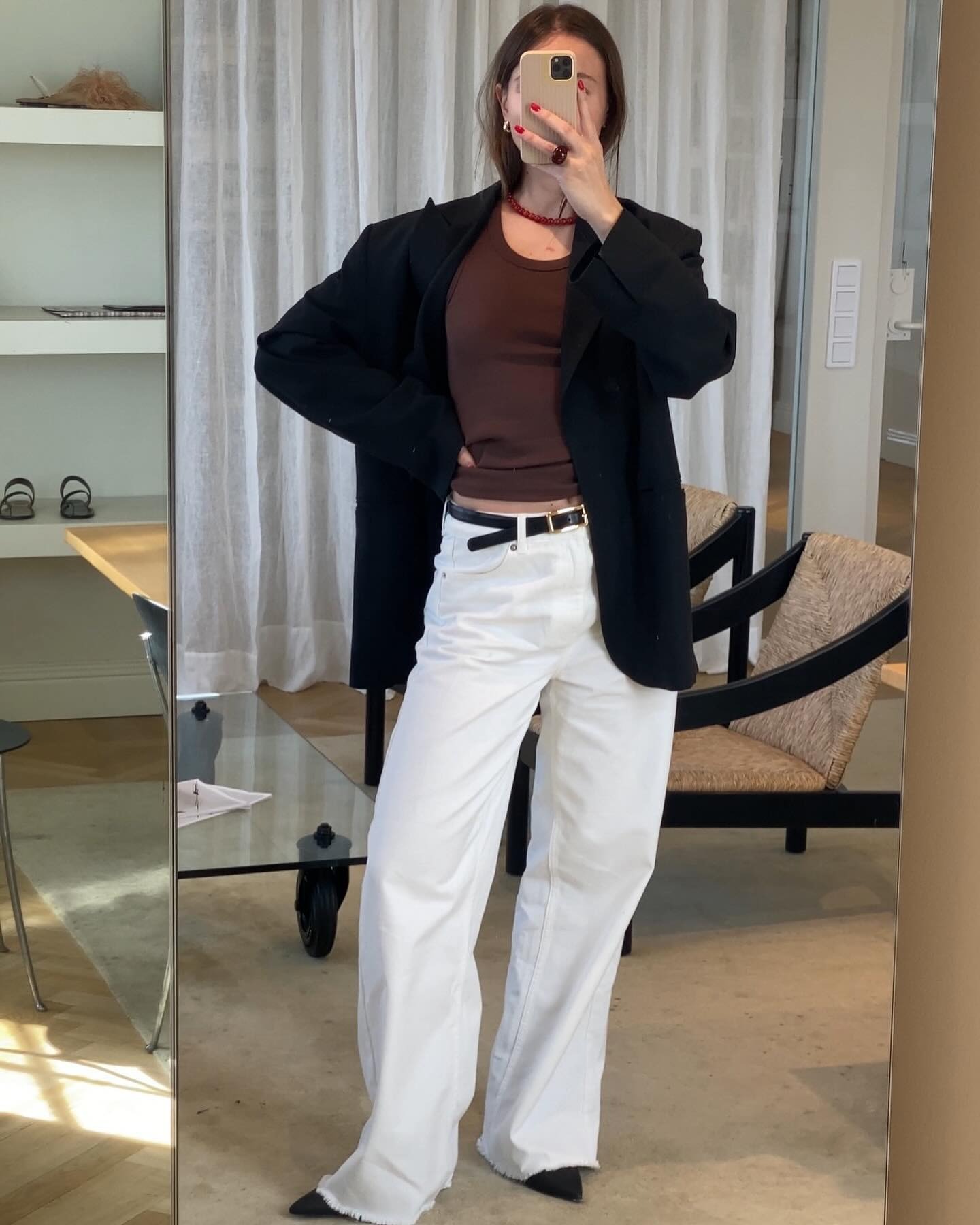 Swedish fashion pr and influencer Johanna Lager poses for a mirror selfie wearing a red beaded necklace, brown T-shirt, oversize black blazer, thin black belt, white wide-leg jeans, and black pointed-toe shoes