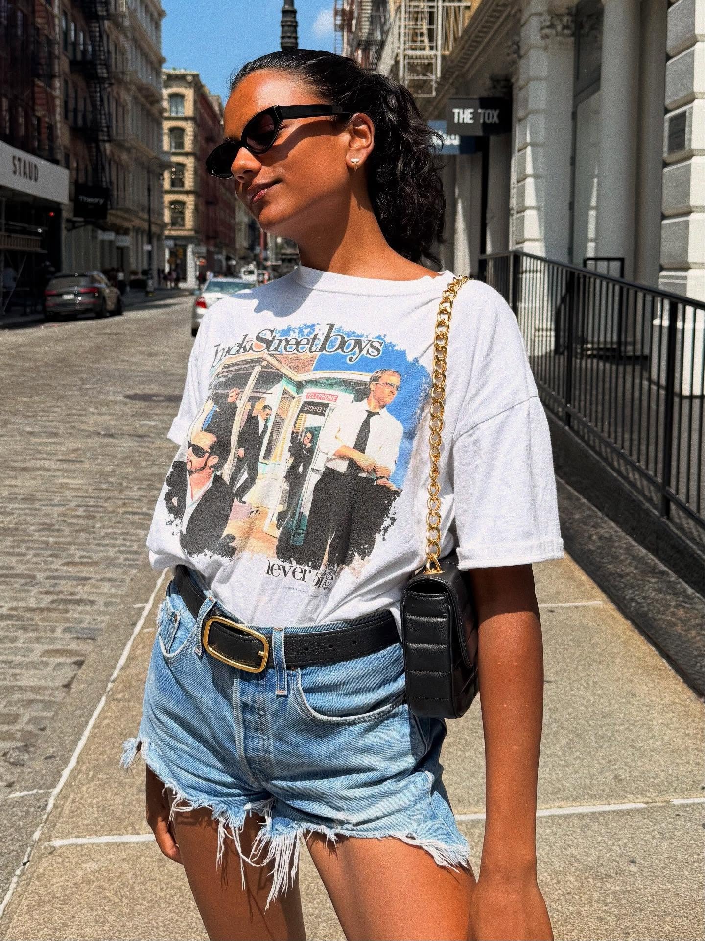 Actor Simone Ashley wears a graphic t-shirt with denim shorts, a leather bag and a black leather belt.