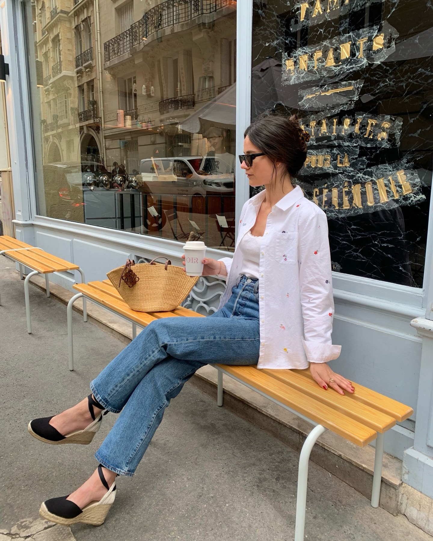 Influencer @leasy_inparis sits on a bench outside of a coffee shop wearing a white shirt, a white vest top and blue high-waisted jeans.