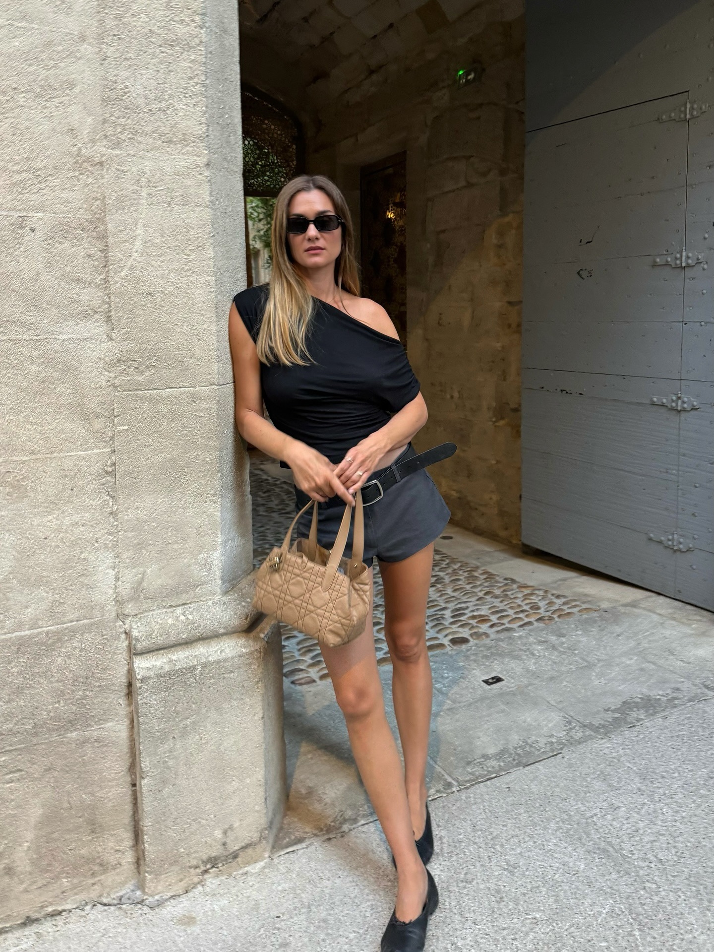 Influencer @annelauremais leans on a door frame wearing grey shorts, a black top and black leather ballet flats.