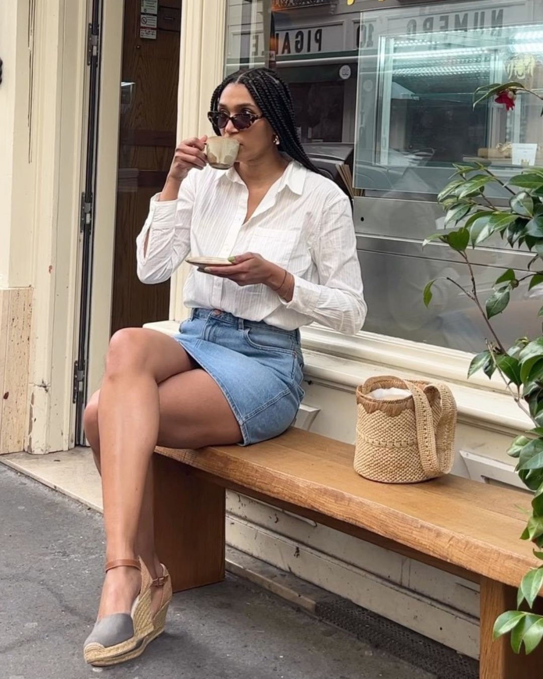 Influencer @lenafarl sits on a bench outside of a coffee shop wearing a white shirt, a denim skirt and espadrille wedges.
