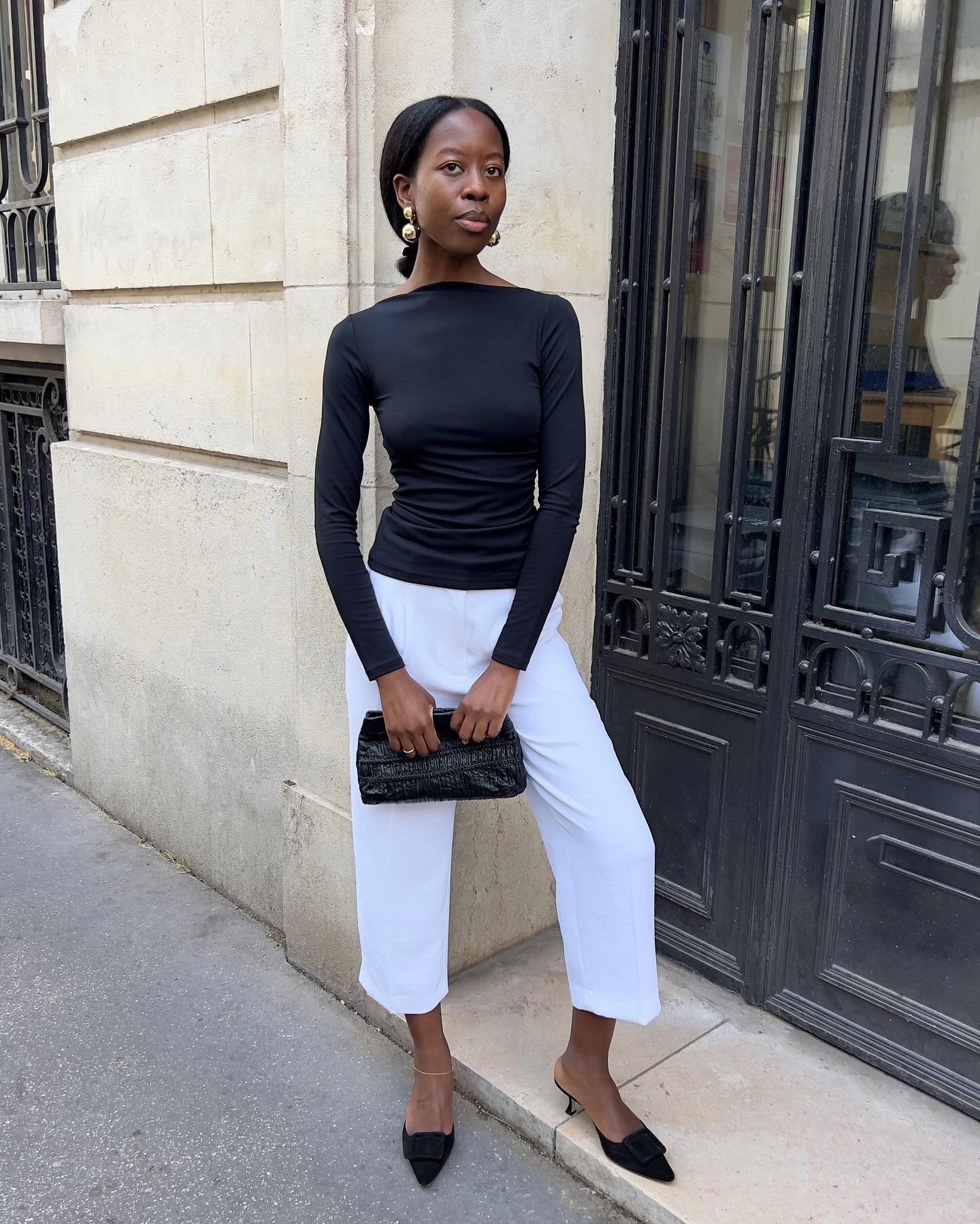 French influencer @sylviemus_ stands on the street in Paris wearing a black boat neck top with white cropped trousers.