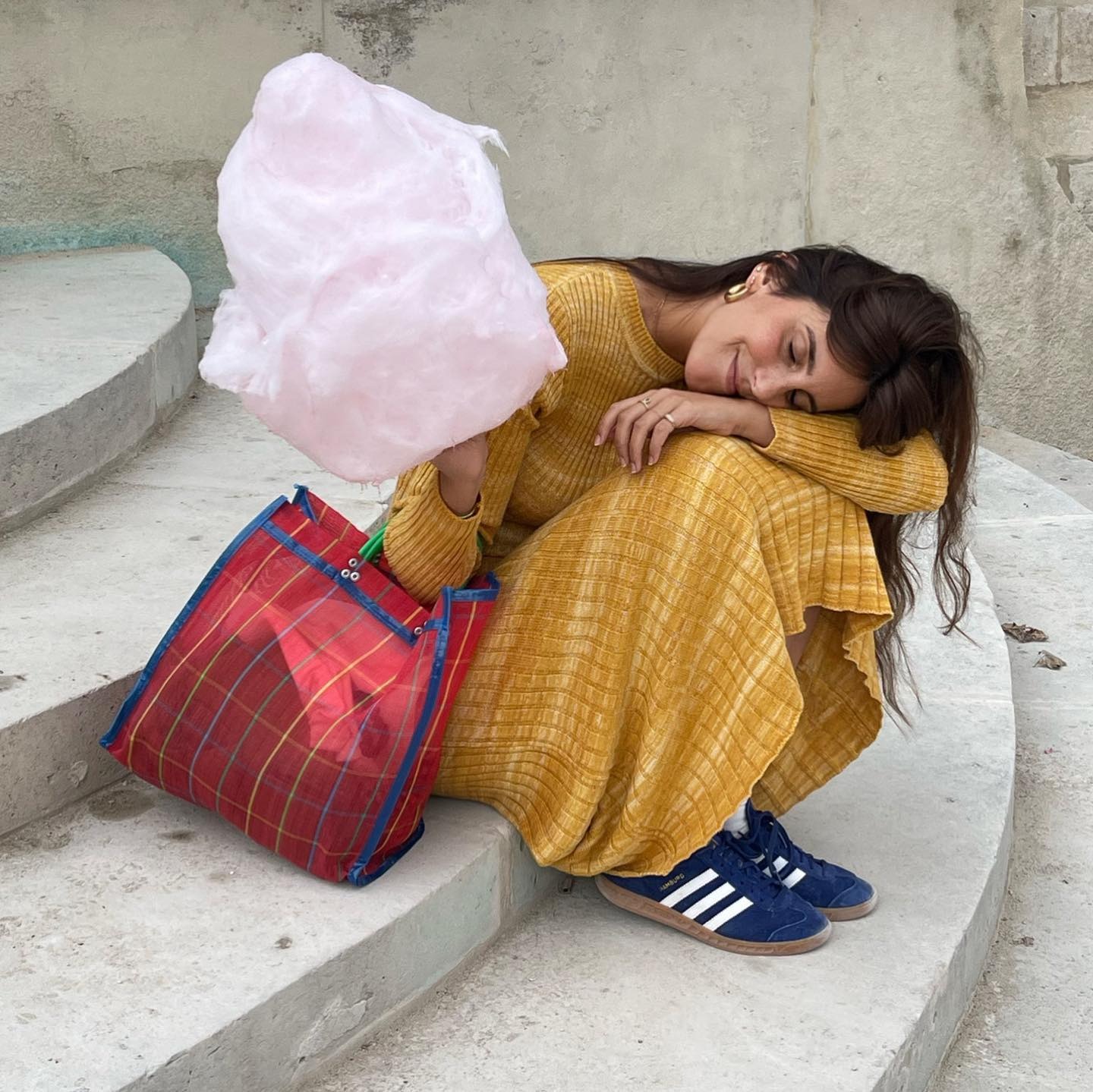 Influencer @dianekari sits down on stairs outside wearing a yellow dress with navy adidas trainers. She holds a stick of candy floss and rests her head on her arm.