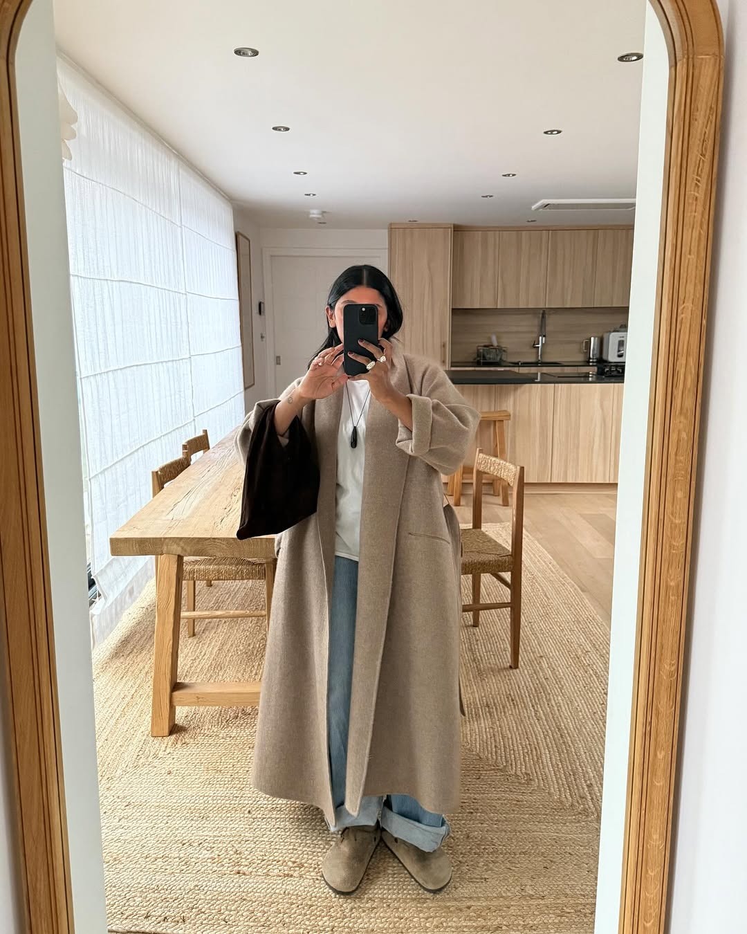 Influencer Monikh wears a long beige coat with cuffed light-wash jeans, a white crewneck, and Birkenstock Boston clogs.