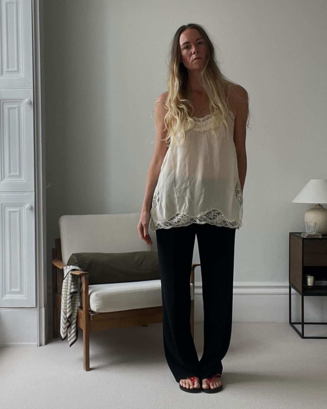 Influencer @samreid___ stands in a bedroom wearing a semi-sheer long-line cream top over black trousers with red flip flops.