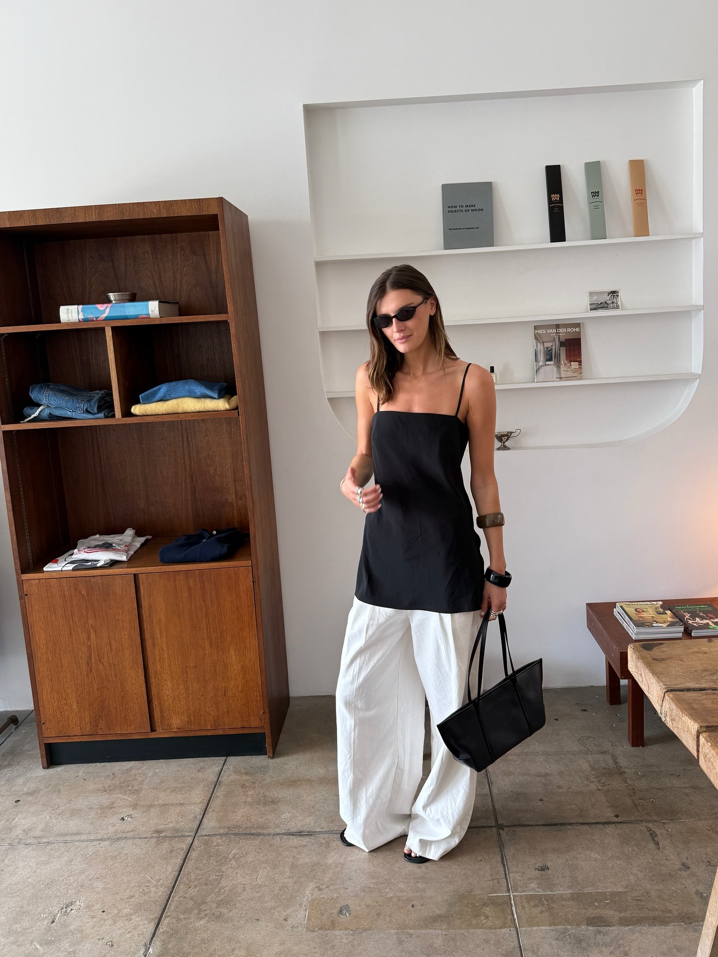 Influencer @cocoschiffer wears a black long top over white wide-leg trousers. She carries an east-west bag and wears chunky bangles on her arm.