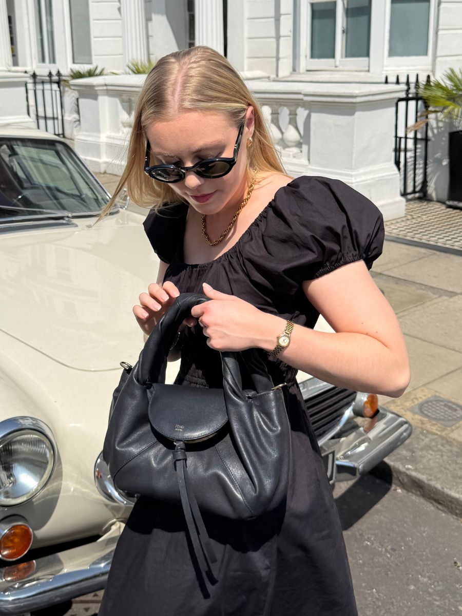 Woman wears black dress, black sunglasses and carries black bag
