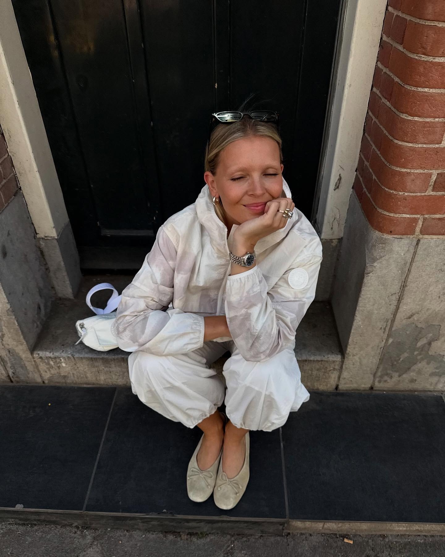 Influencer @holliemercedes sits outside on a doorstep wearing a white and lilac windbreaker, suede ballet ballet flats. She rests her face in her hand and smiles with her eyes closed.