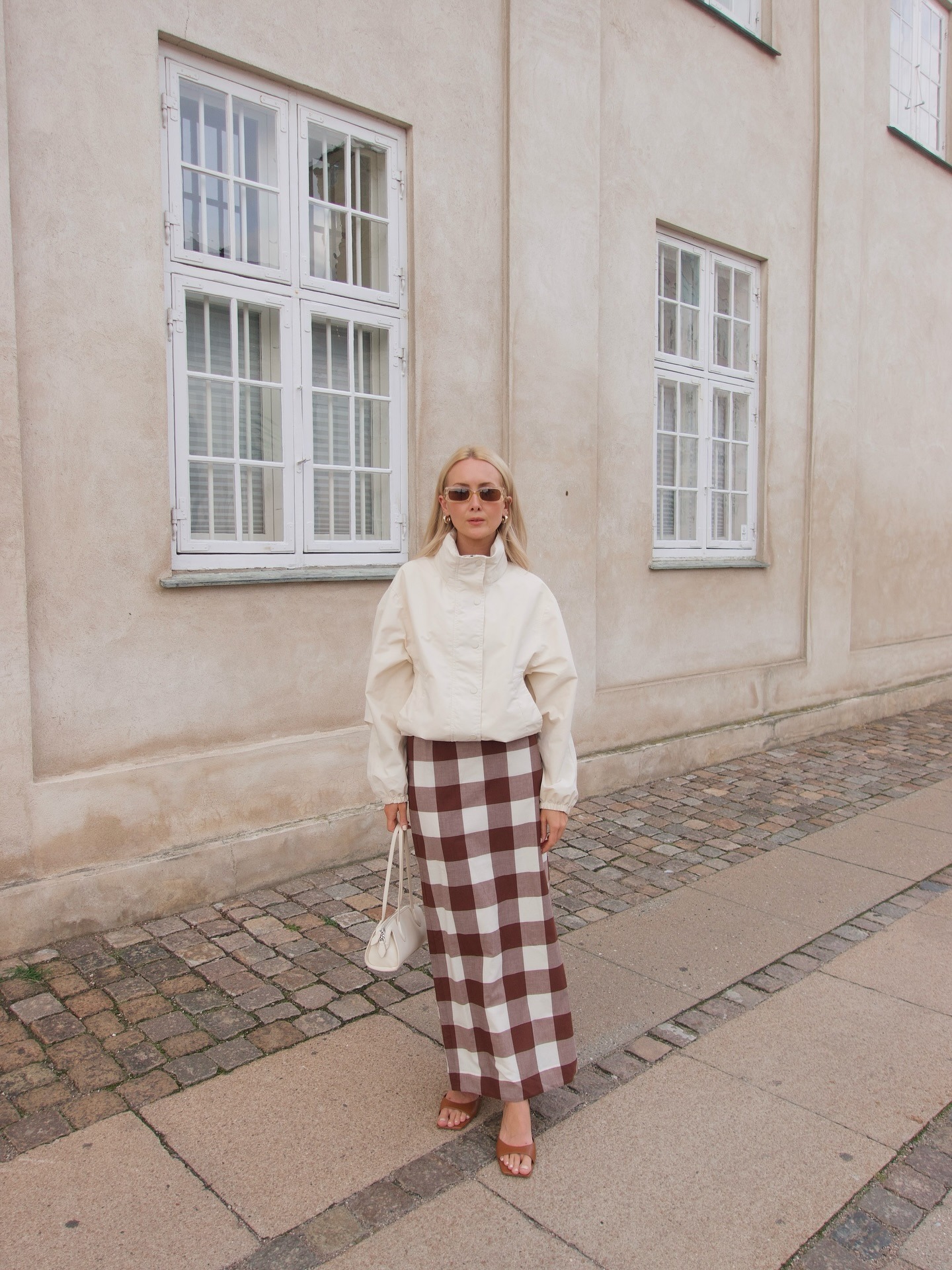 Influencer @chloekathbutler stands outside a light cream building wearing a white windbreaker jacket with a gingham maxi pencil skirt. She holds a while east-west bag in her hands and wears blight brown heeled sandals and brown sunglasses.