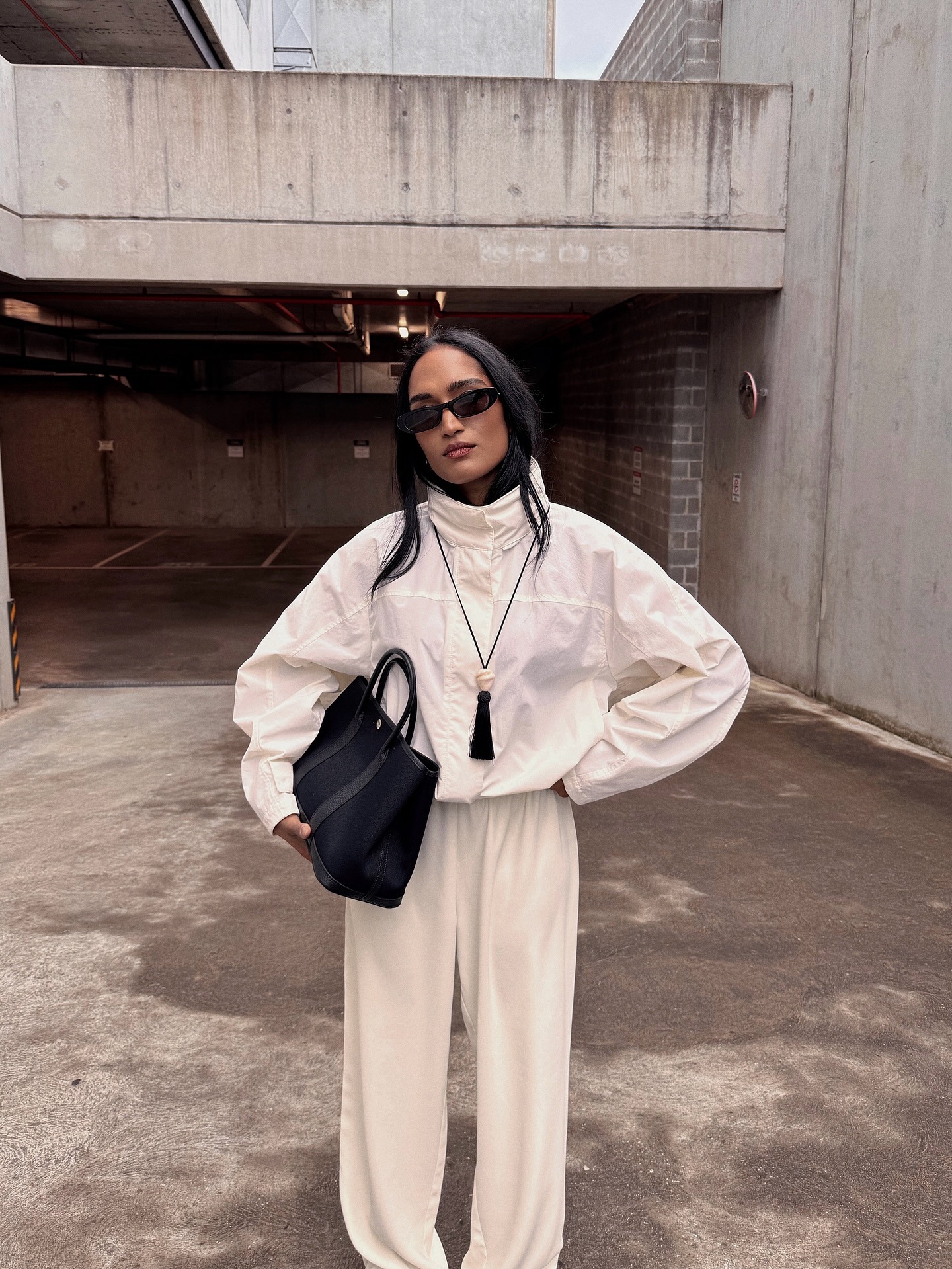 Influencer @divrav stands outside wearing a white windbreaker jacket from Uniqlo with white trousers and a suede bag. She wears a long tassel necklace and black sunglasses and stands with one hand on her hip.
