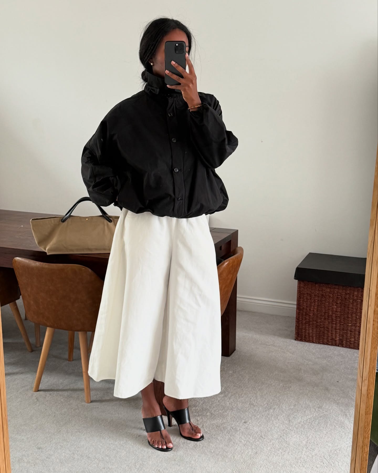 Influencer@nlmarilyn takes a mirror selfie indoors, wearing a black windbreaker jacket, fastened to the top with a white skirt and black heels.