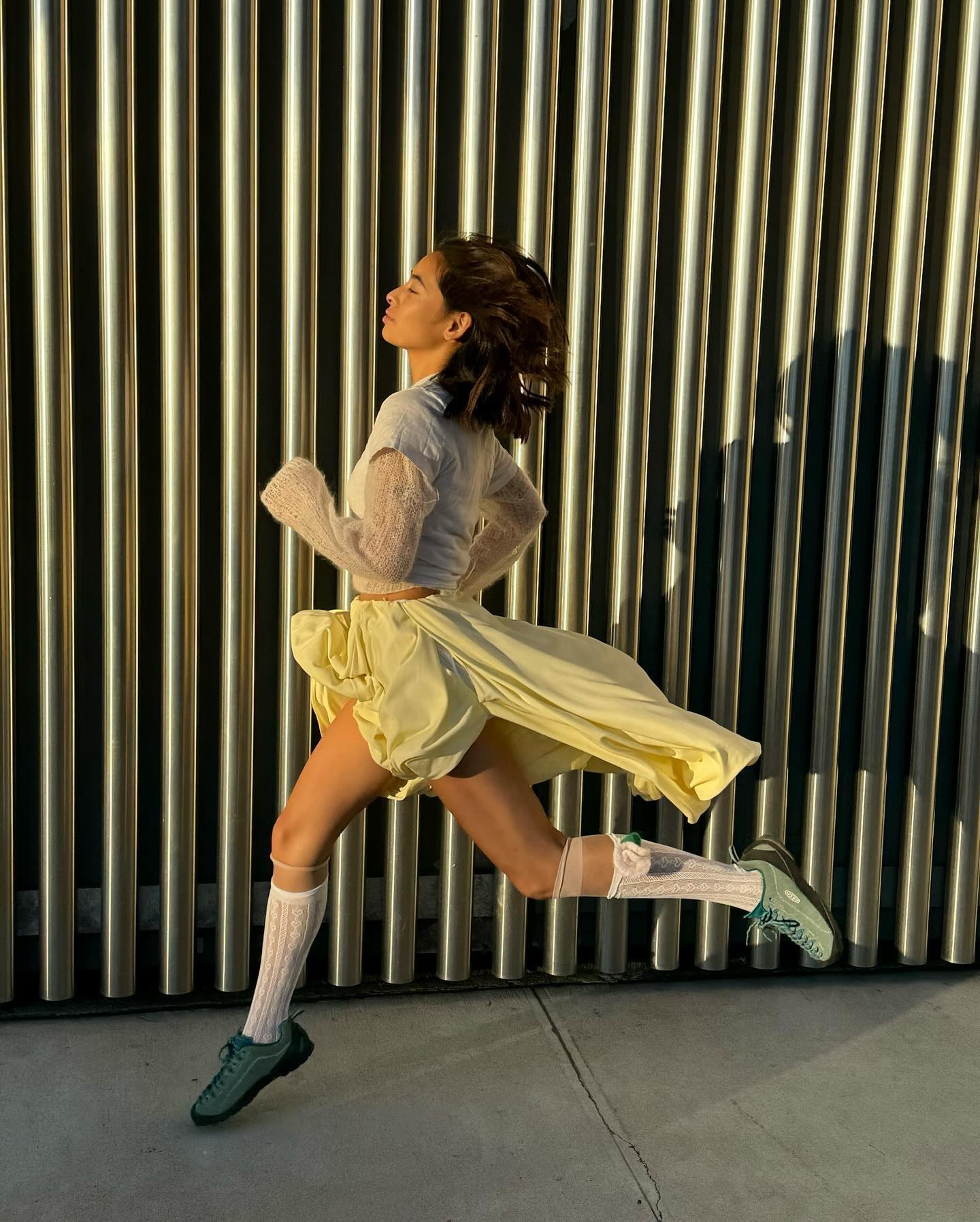 Influencer @himichelleli jumps through the air wearing green Keen trainers with a white skirt and grey top.