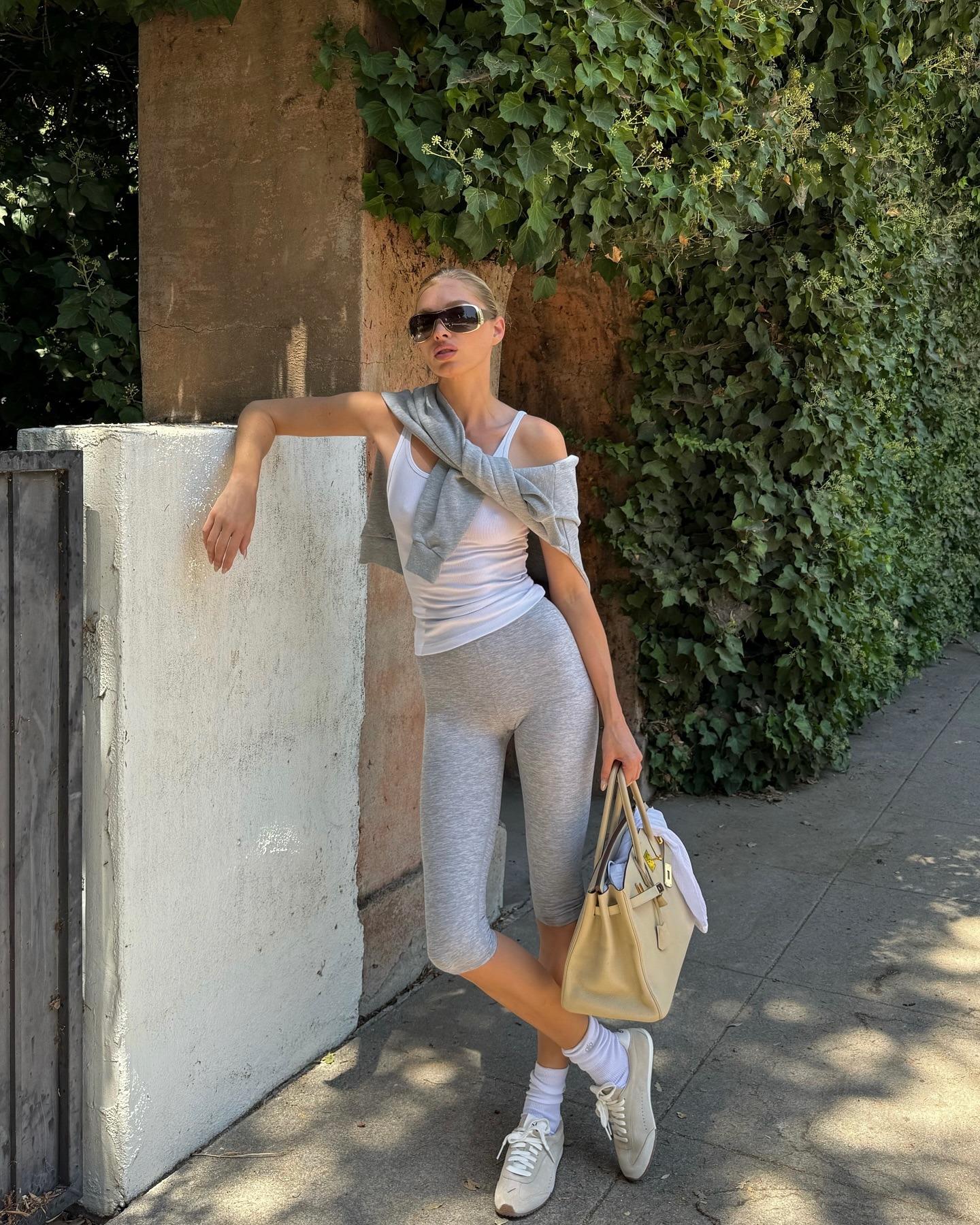 Model Elsa Hosk leans on a fence outside wearing grey capri pants with a white vest top, a Hermes bag and grey trainers styled with white socks.
