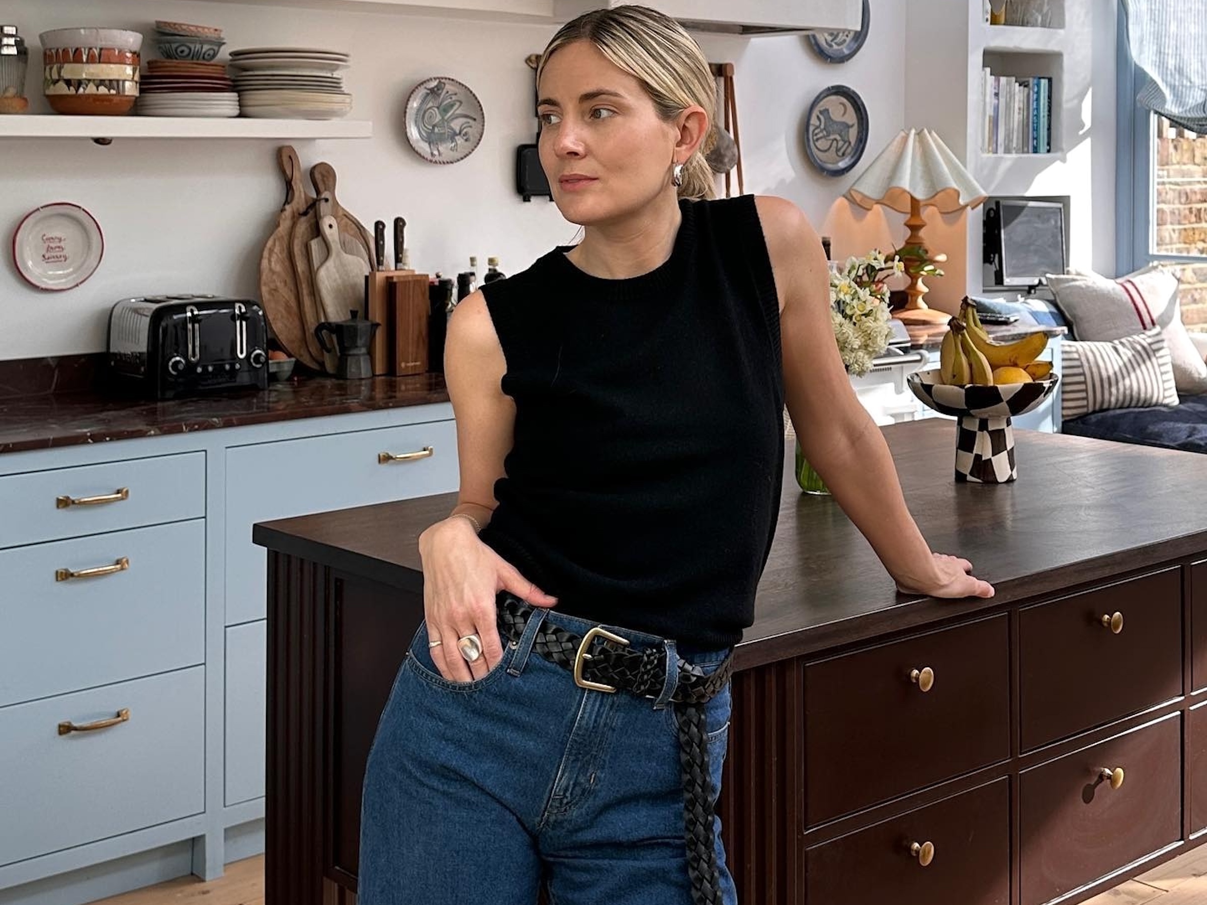 British style influencer Lucy Williams poses in her chic London kitchen with light blue cabinets wearing a simple black tank top, woven black belt, and high-waisted jeans
