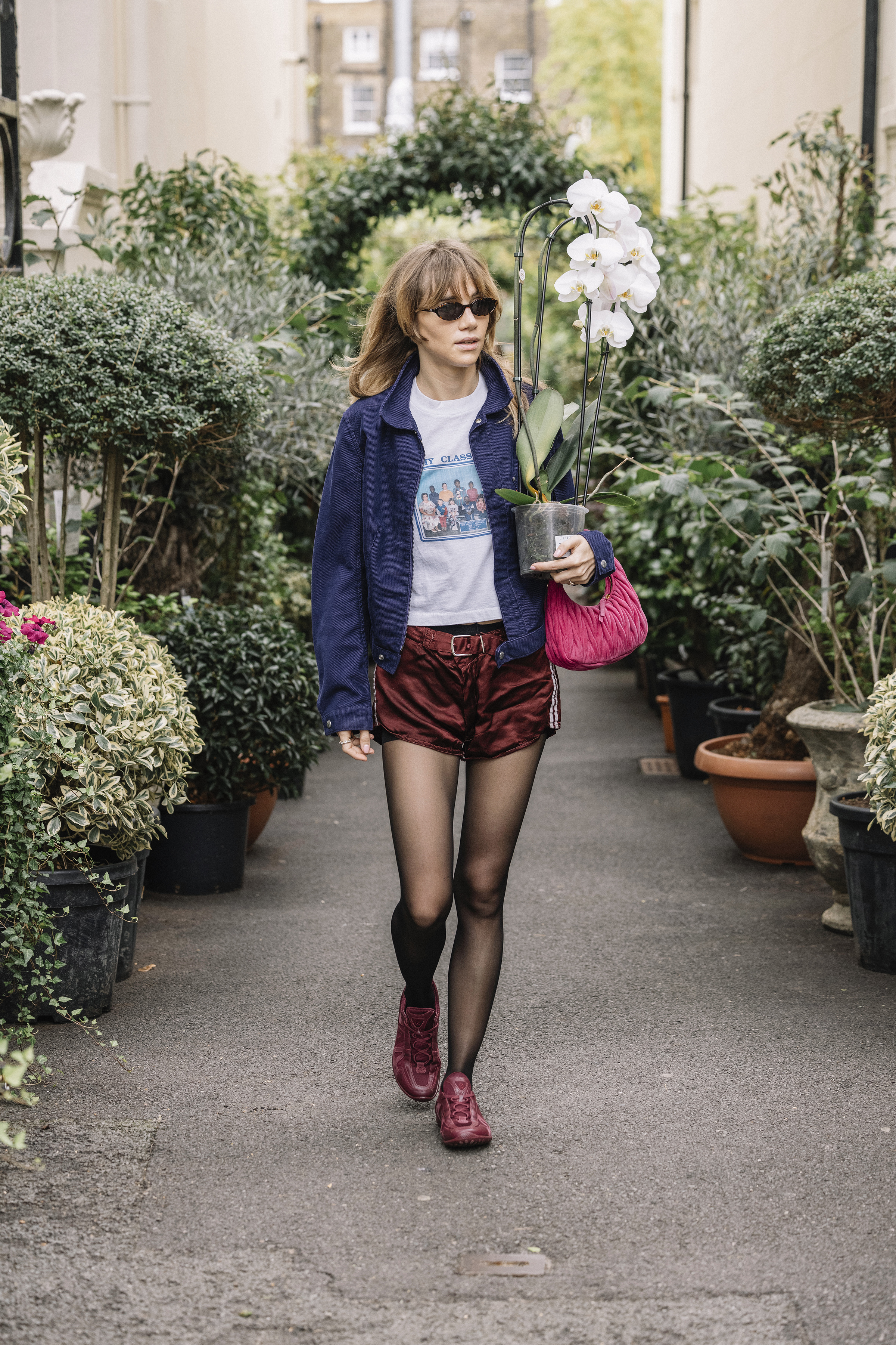 Suki Waterhouse wearing a navy jacket, white T-shirt, and burgundy shorts and sneakers while carrying a plant in London