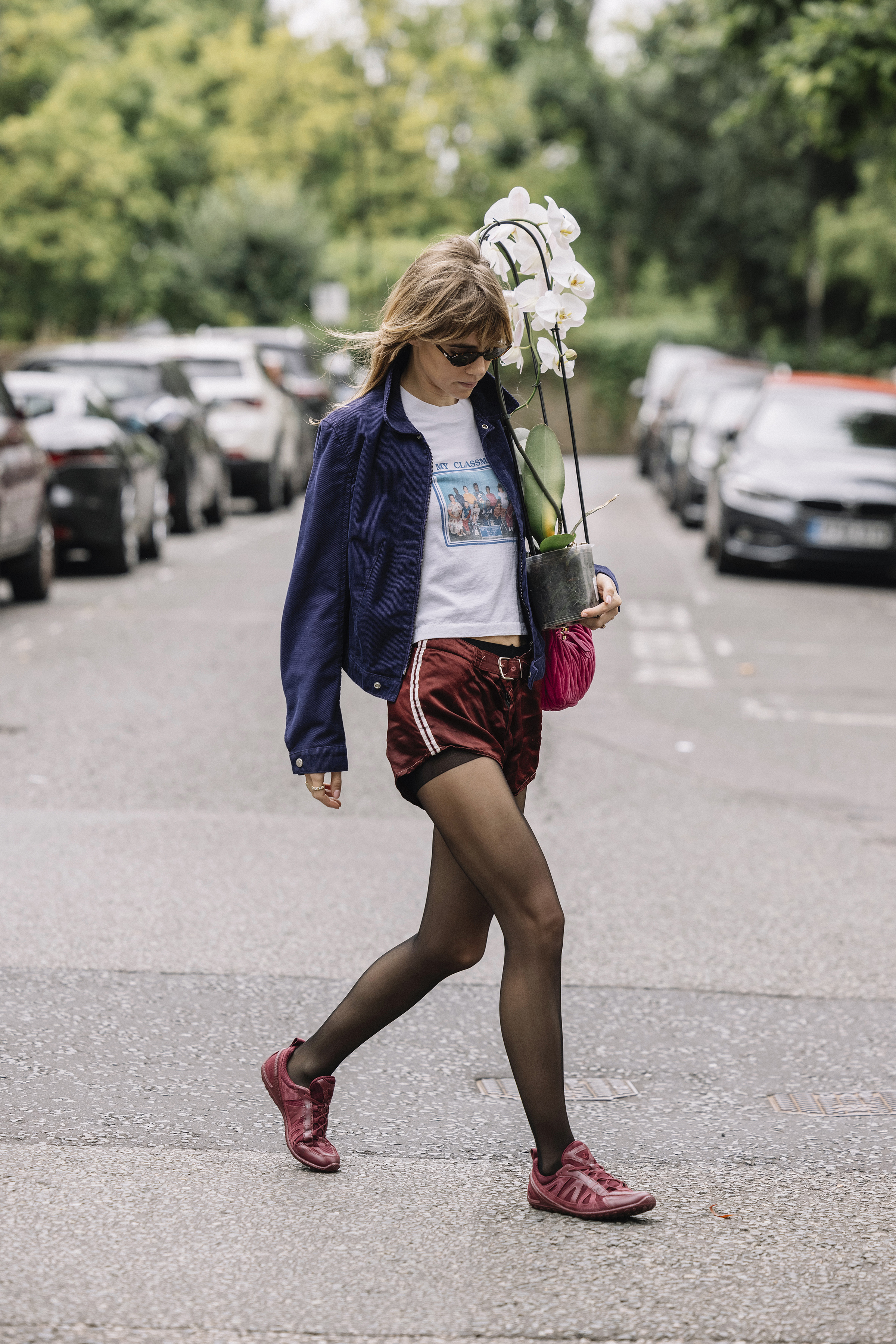 Suki Waterhouse wearing a navy jacket, white T-shirt, and burgundy shorts and sneakers while carrying a plant in London