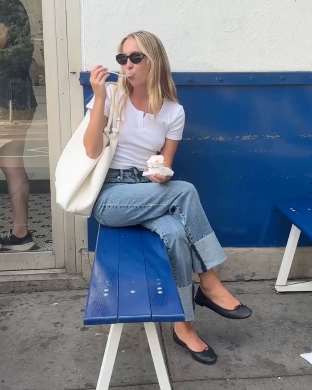 Model Lila Moss sits on a bench outside wearing ice cream, wearing a white t-shirt, blue jeans, ballet flats and a white handbag.