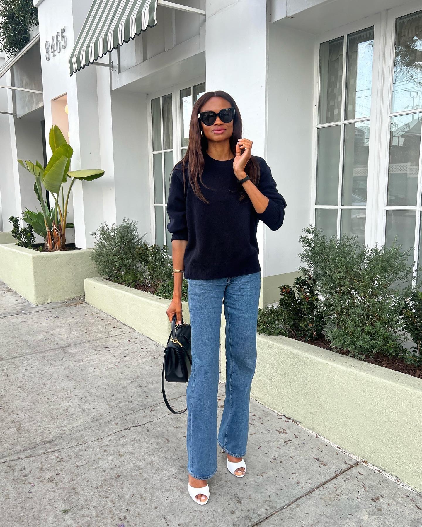 Influencer @lornasymphony stands outside wears a navy jumper with mid-wash blue jeans and white open-toe heels.