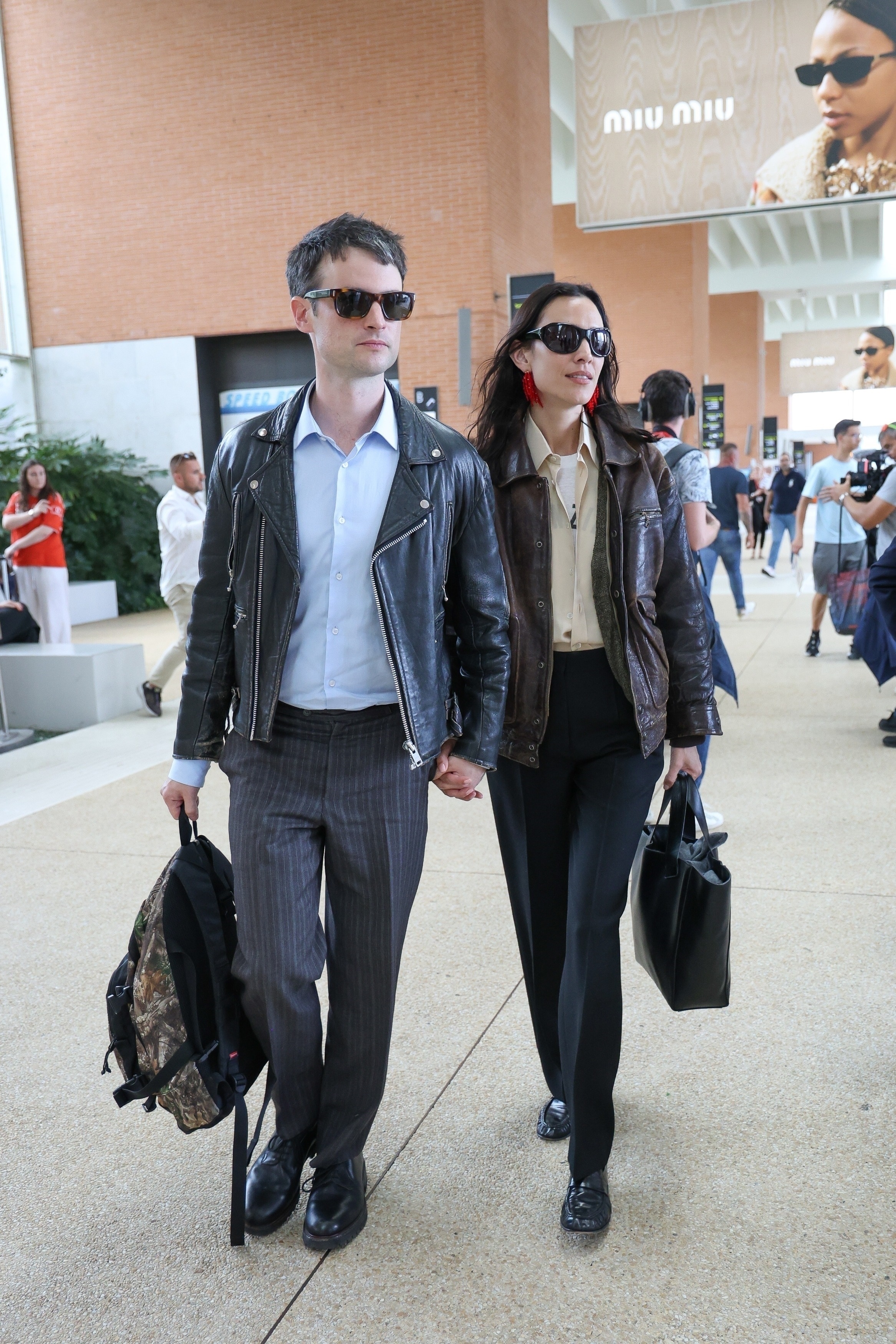 Alexa Chung and Tom Sturidge walk through Venice airport holding hands. Alexa carries a black bag in her hands and wears a brown leather jacket, a yellow shirt, black trousers and black loafers.