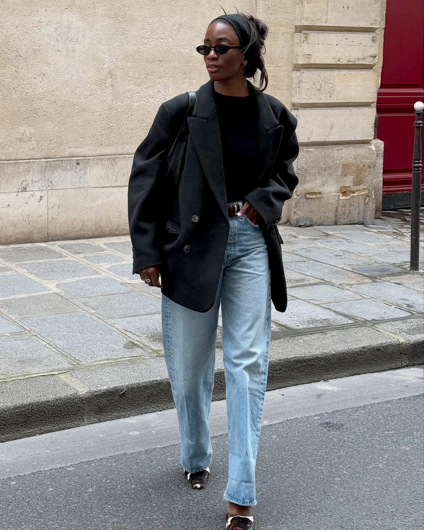 British style influencer Liv Madeline crosses a street inParis wearing a black headband, black oval sunglasses, an oversize black blazer jacket, black top, black belt, light-wash barrel jeans, and cow-print mule heels
