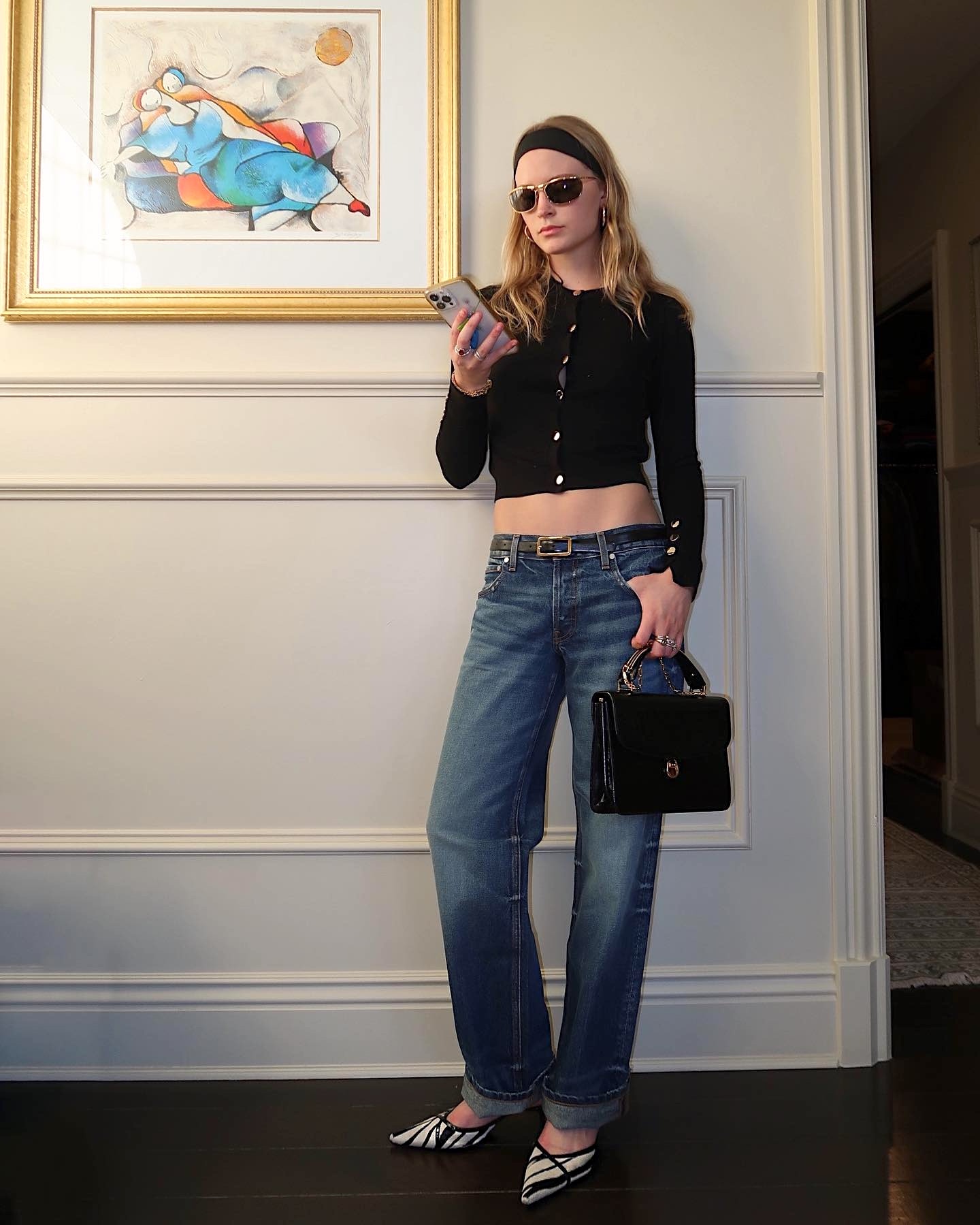 style influencer Lauren Elson poses while looking at her cellphone wearing a black headband, metal-rim sunglasses, a cropped black cardigan, black skinny belt, cuffed jeans, and zebra-print pointed-toe heels