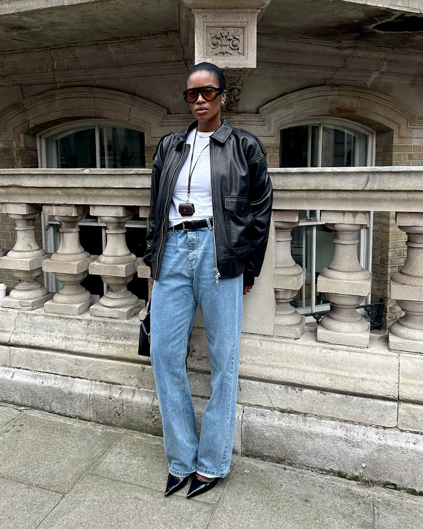 British style influencer Marilyn Nwawulor-Kazemaks poses in London wearing a fall 2025 outfit with a black leather moto jacket, cord lanyard necklace, oversized black aviator sunglasses, a white T-shirt, black belt, relaxed jeans, and black pointed-toe shoes