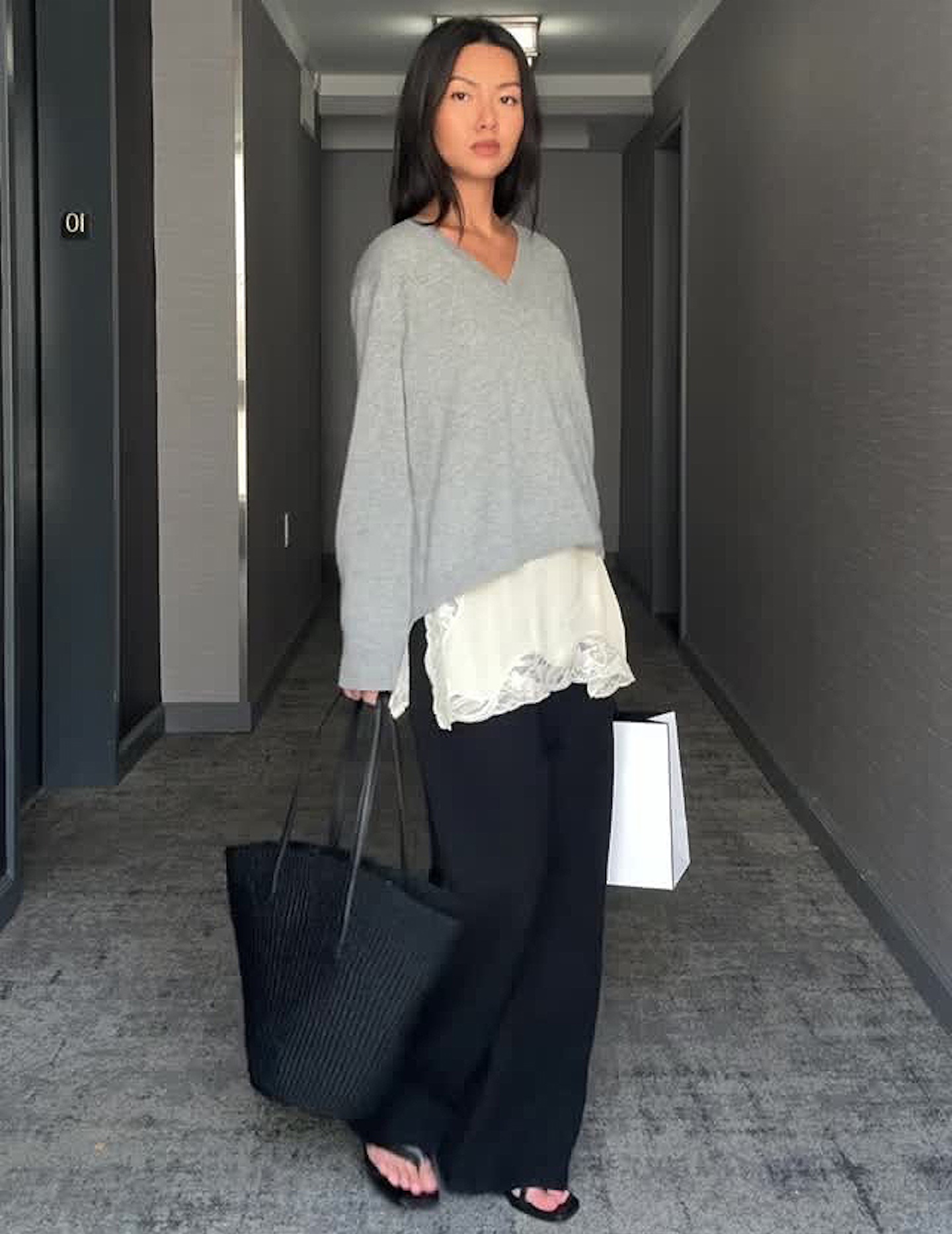 NYC style influencer Mimi Nguyen poses in a chic fall 2025 outfit with a light gray v-neck sweater layered over a white lace-hem silk top, wide-leg black pants, a woven black tote bag, and black flip-flop sandals