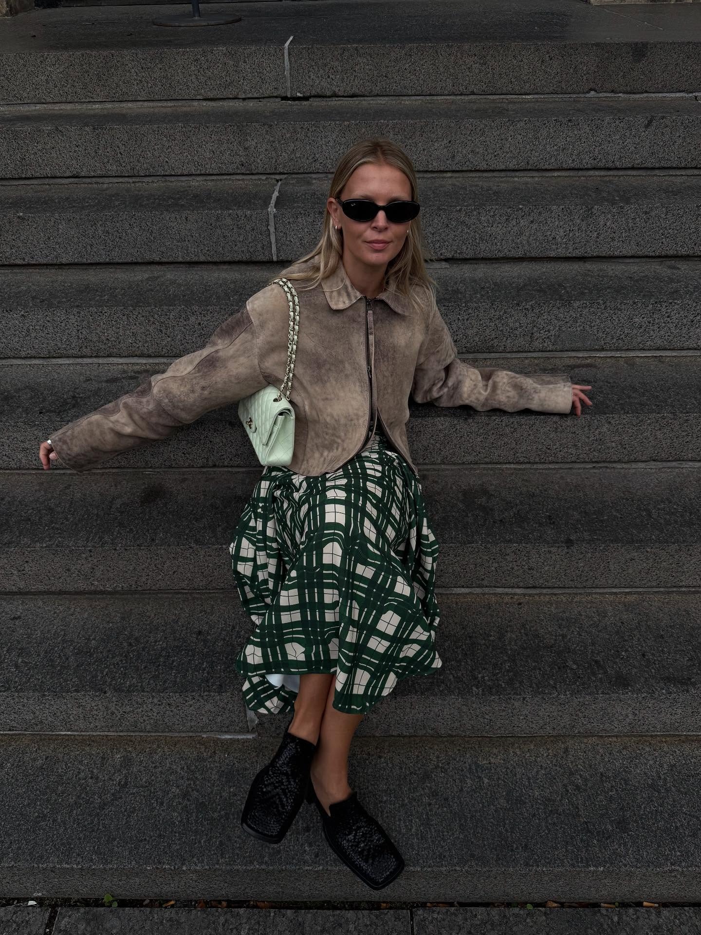 Influencer @holliemercedes wears a checked skirt with a light brown suede jacket and black square-toe boat shoes.