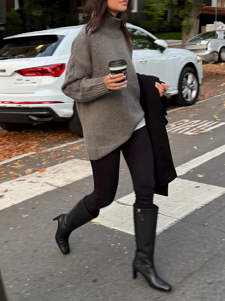 NYC style influencer Mimi Nguyen walks across a crosswalk with a coffee in hand wearing an oversize neutral brown turtleneck sweater, black leggings, and black heeled knee-high boots