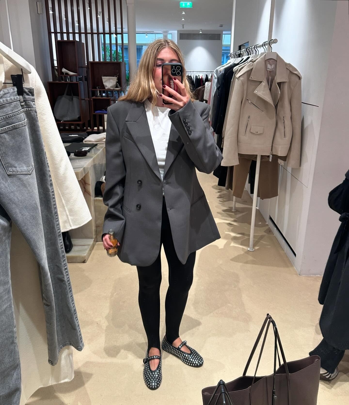 Danish style influencer and fashion creative Sidsel Alling poses for a mirror selfie wearing a gray oversize blazer, white T-shirt, black stirrup leggings, and black studded Alaia Mary Jane flats
