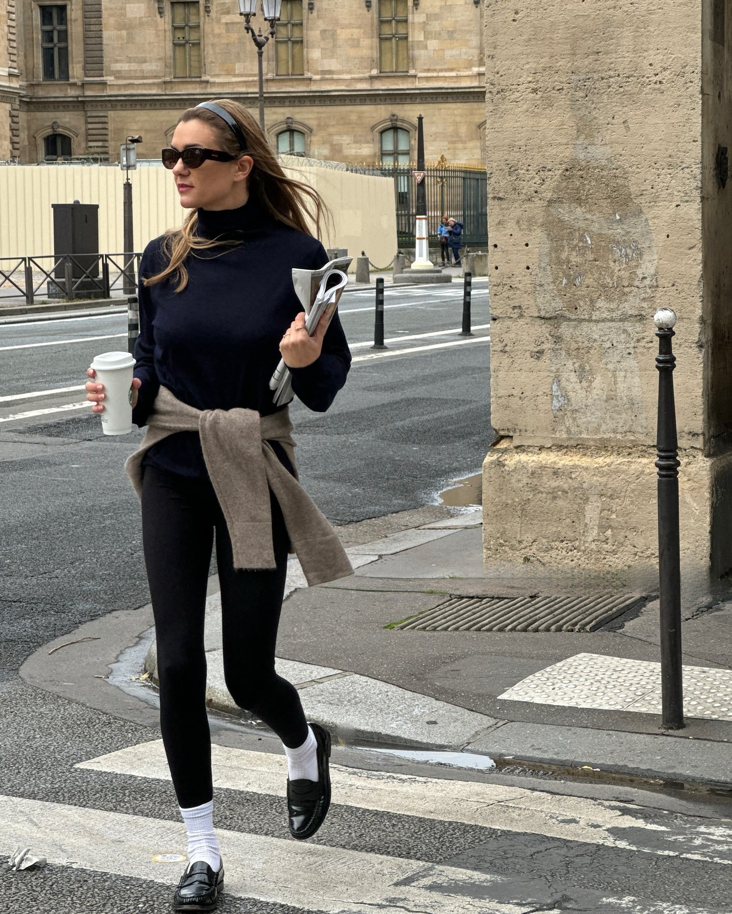 French influencer walks across a Paris crosswalk holding a coffee wearing black sunglasses, a black turtleneck top, a tan sweater tied around her waist, black leggings, white ankle socks, and black loafers