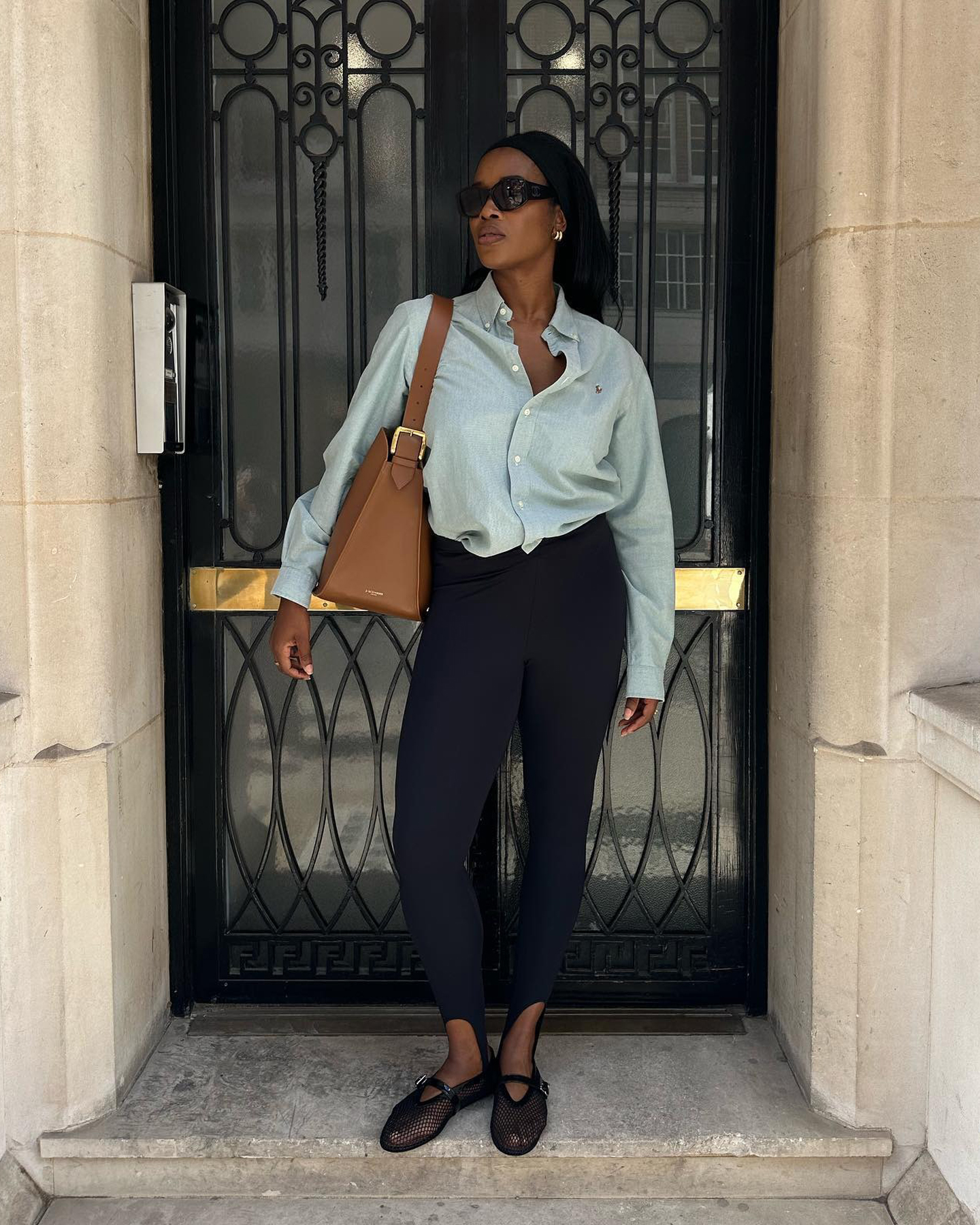British fashion influencer Marilyn Nwawulor-Kazemaks poses in a doorway in London wearing oversize black sunglasses, a light-wash denim button-down shirt, tan shoulder bag, black stirrup leggings, and Alaia mesh Mary Jane flats