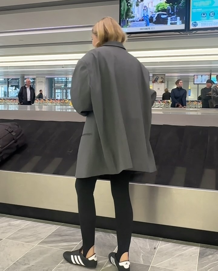 Danish style influencer and fashion creative Sidsel Alling waits at an airport luggage carousel wearing an oversize gray blazer, black stirrup leggings, and black Adidas sneakers