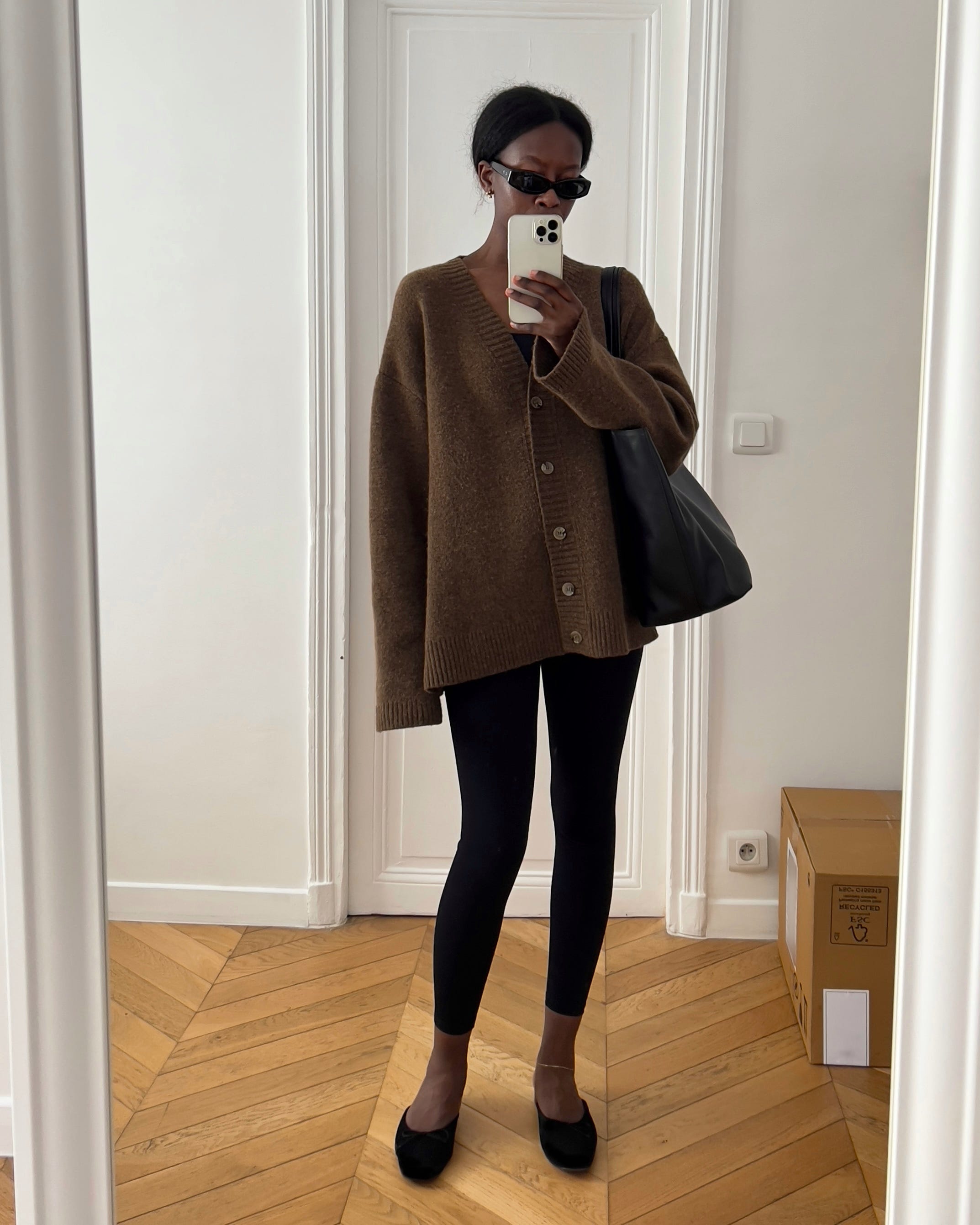 Paris-based fashion influencer and stylist Sylvie Mus poses for a mirror selfie wearing black sunglasses, a brown oversize cardigan sweater, black tote bag, black cropped leggings, and black suede mule flats