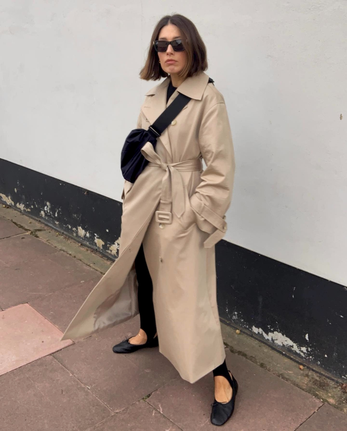 British fashion influencer Lucy Alston poses on a London sidewalk wearing a long belted trench coat, a black crossbody bag, black stirrup leggings, and black ballet flats