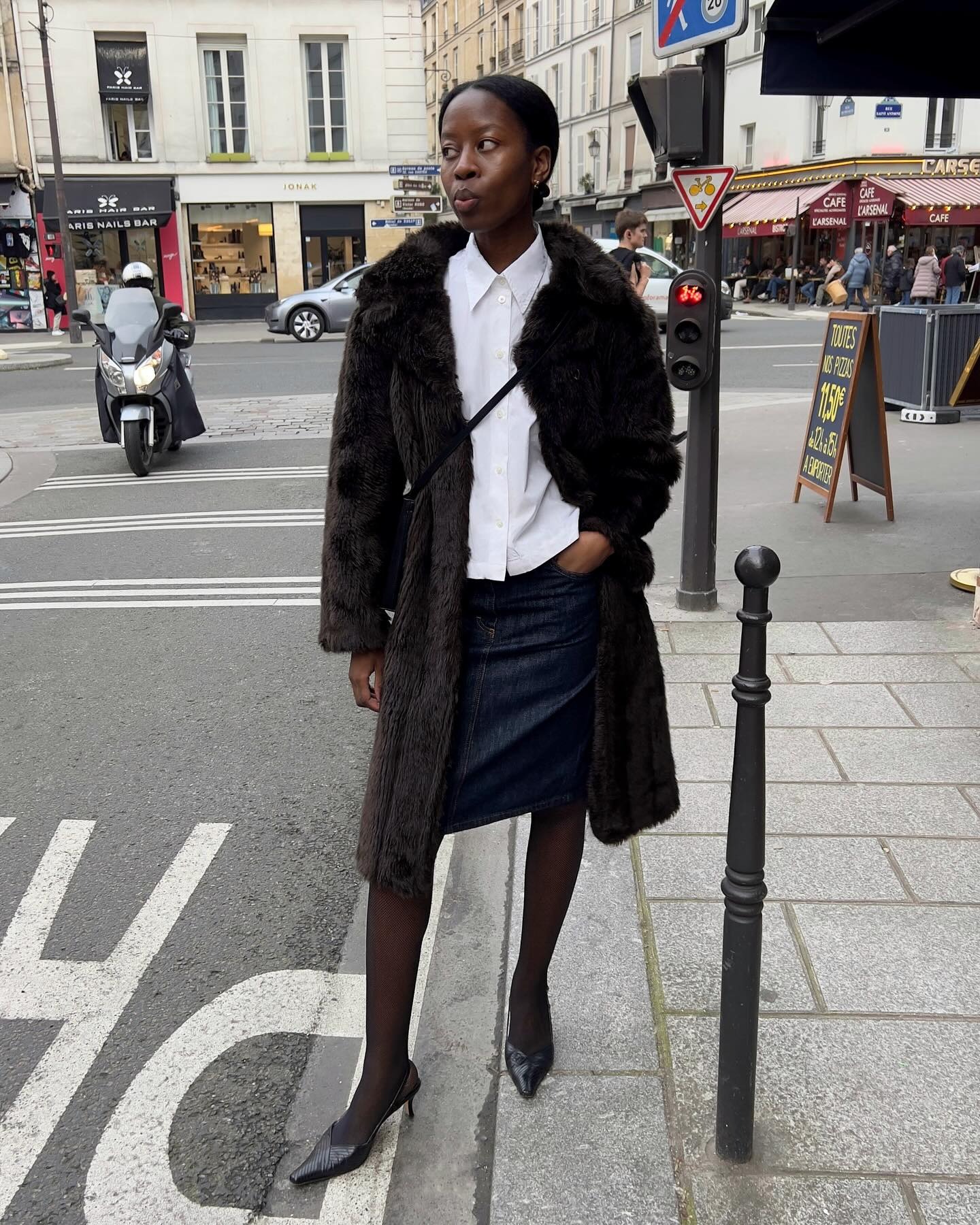 Sylvie wears a brown fur coat, white button-down, denim skirt, sheer tights, and slingbacks.