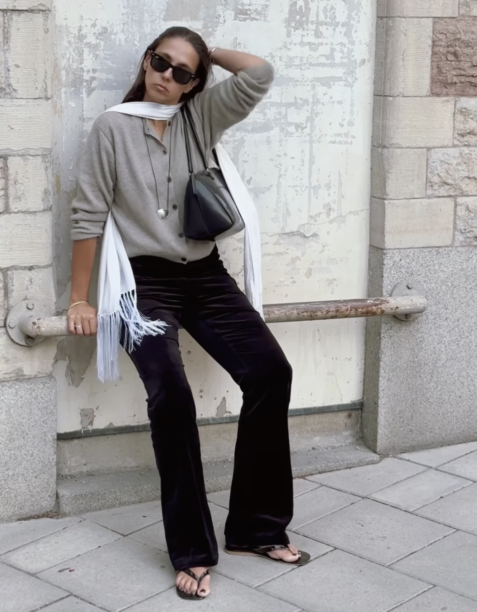 Style influencer Hanna MW poses in black sunglasses, a white scarf, a gray cardigan, a cord necklace with a silver pendant, a black shoulder bag, brown velvet pants, and flip-flip sandals