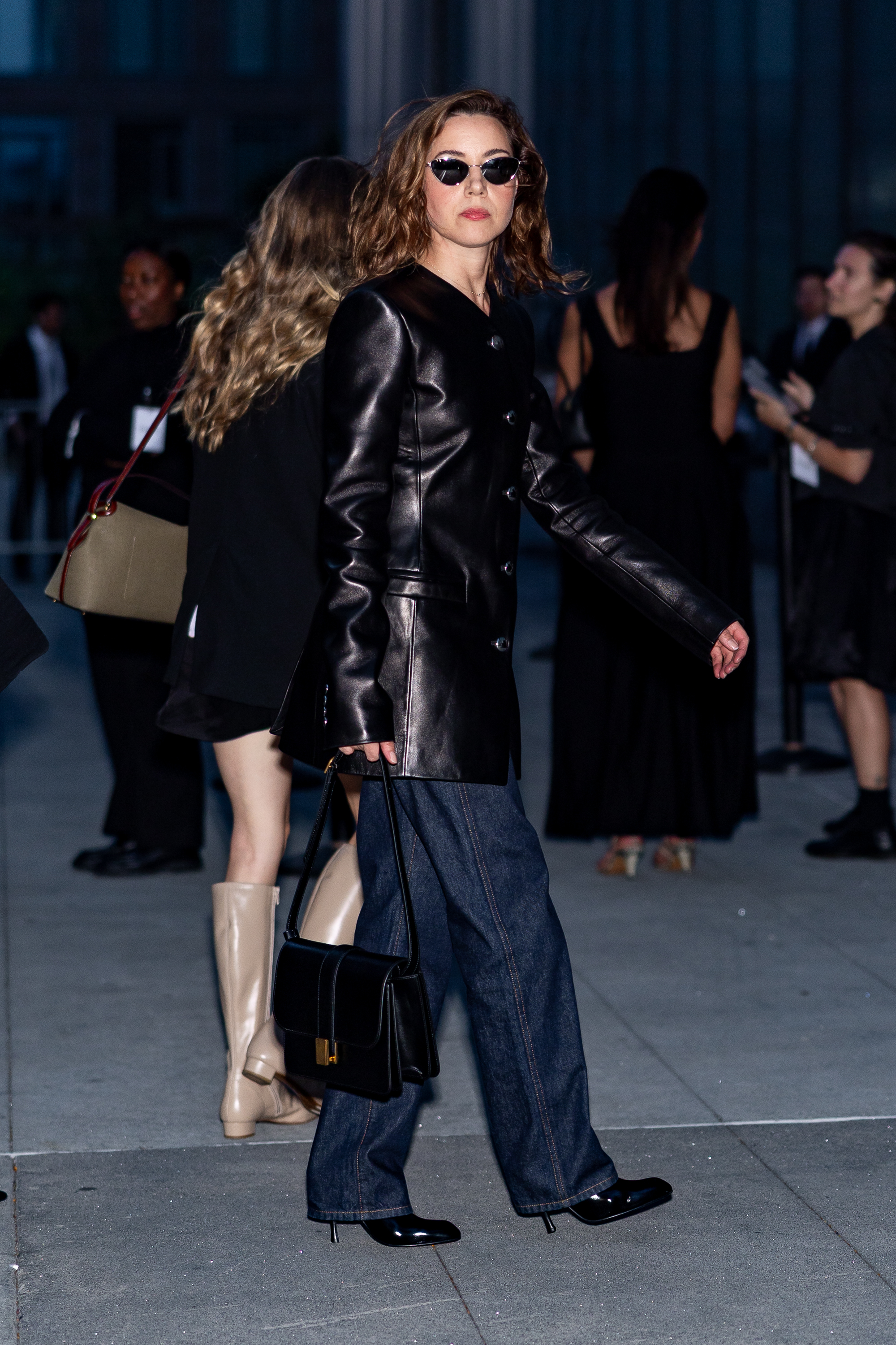 Aubrey Plazer wears a single breasted blazer in black leather with indigo jeans and patent leather boots. She carries a black leather handbag and wears dark black sunglasses.