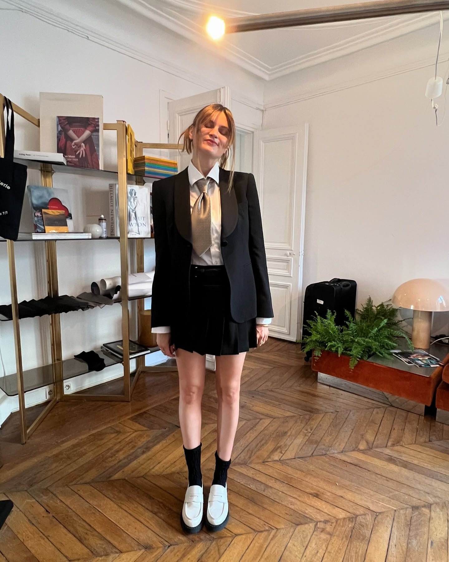 French writer, editor, and creative Eugenie Trochu poses in a Paris apartment with chevron wood floors wearing a tuxedo blazer, white button-down shirt, satin tie, black pleated skirt, black ankle socks, white chunky loafers