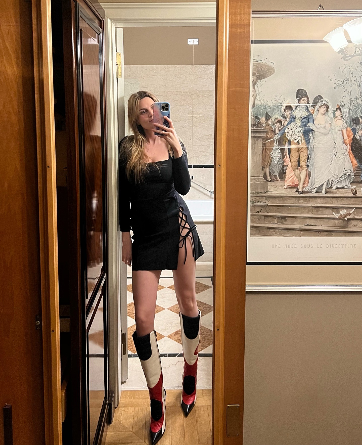 French writer, editor, and creative Eugenie Trochu poses in a hotel in Milan, Italy wearing a black long-sleeved dress with a lace-up hem and leather colorblock knee-high boots