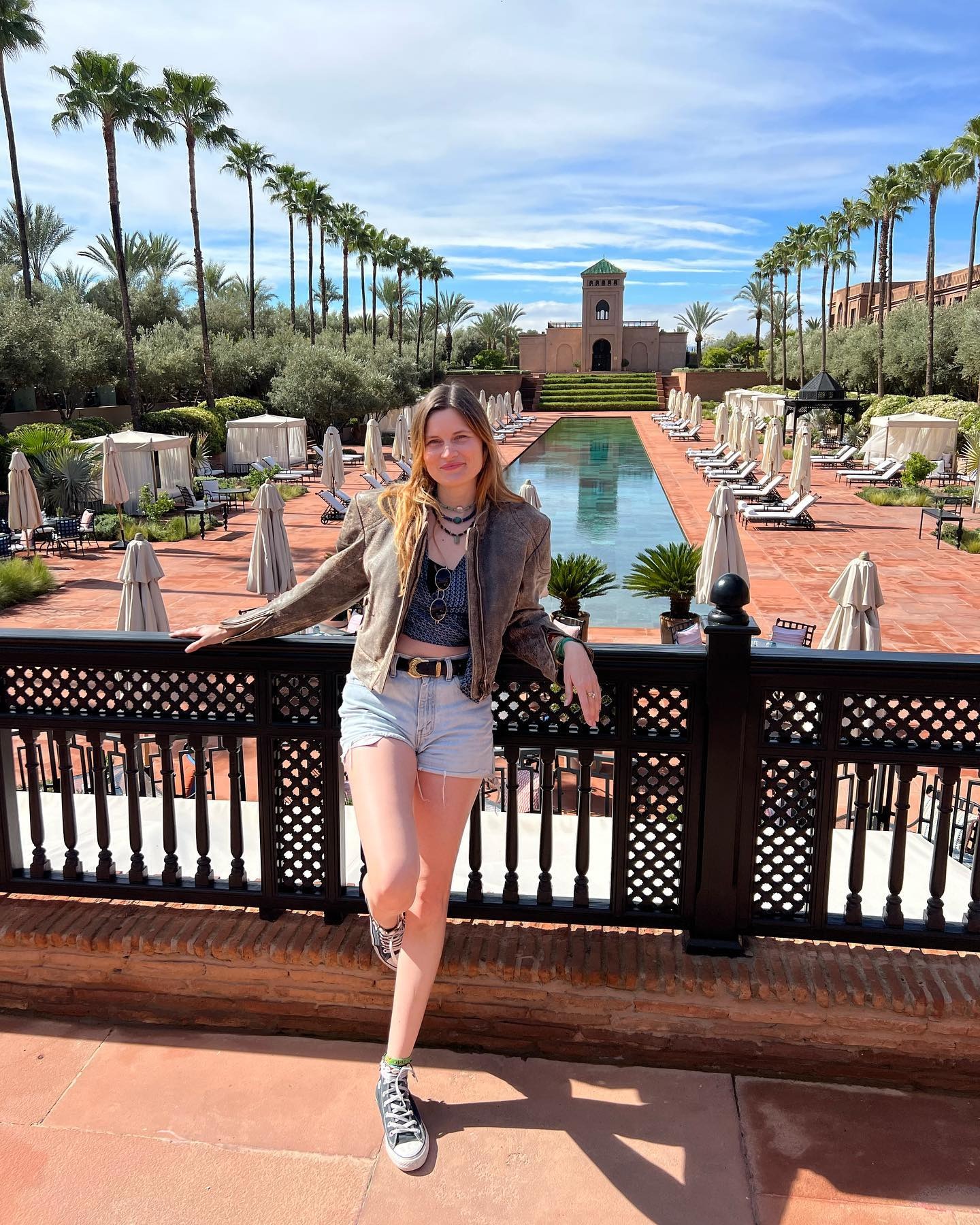 French editor, writer and creative Eugenie Trochu poses in Marrakech wearing a cropped brown leather jacket crop top, western-inspired belt, cut-off denim shorts, and Converse sneakers