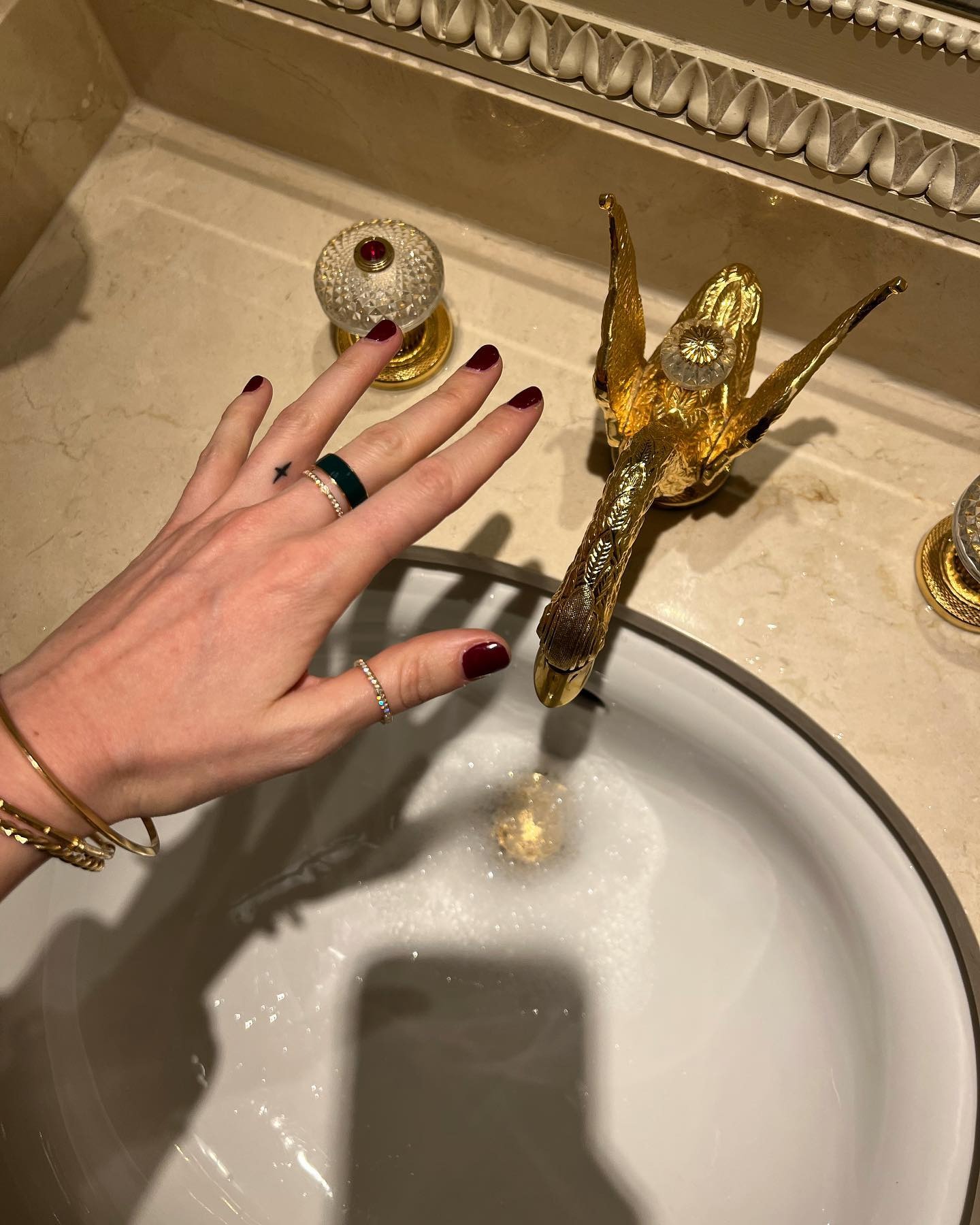 French editor, writer and creative Eugenie Trochu poses her hand over an ornate gold sink at The Ritz in Paris with burgundy nails, stacked rings and yellow-gold bracelets