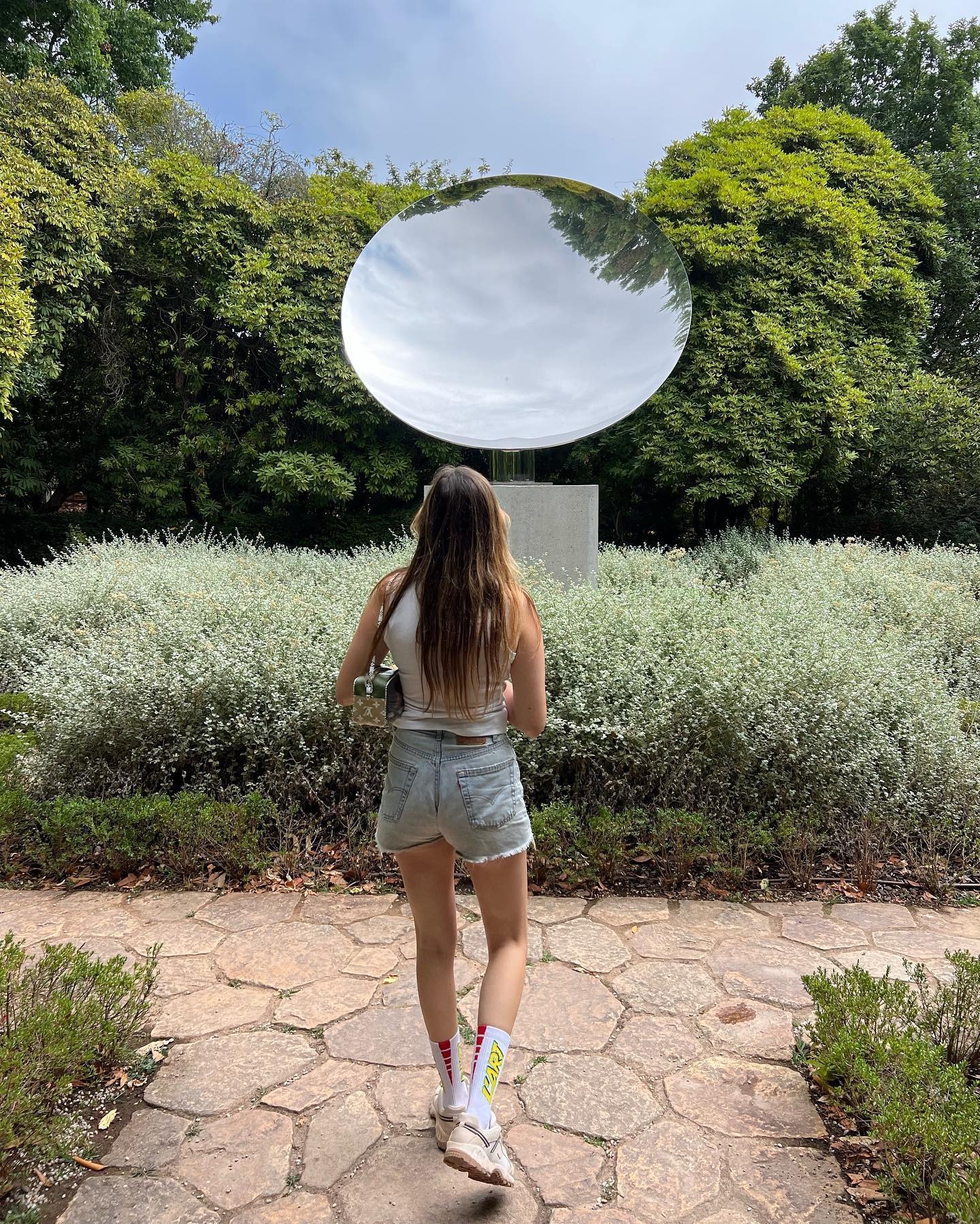 French writer, editor, and creative Eugenie Trochu poses in front of a sculpture in a garden in Porto, Portugal wearing a white tank top, a mini Louis Vuitton shoulder bag, light-wash cut-ff denim shorts, white graphic ankle socks, and sneakers